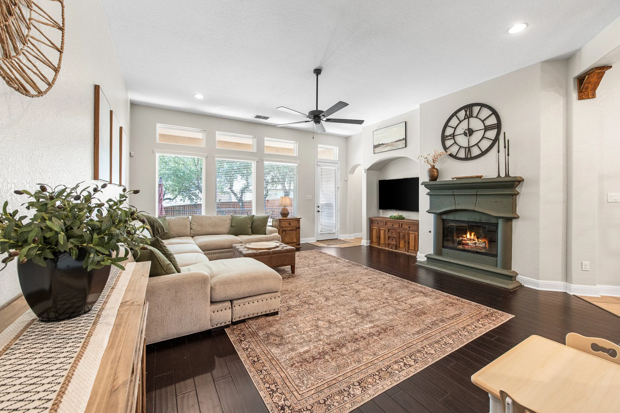 Cozy living room with beige sectional sofa, area rug, fireplace, wall clock, TV, wooden furniture, and large windows letting in natural light.