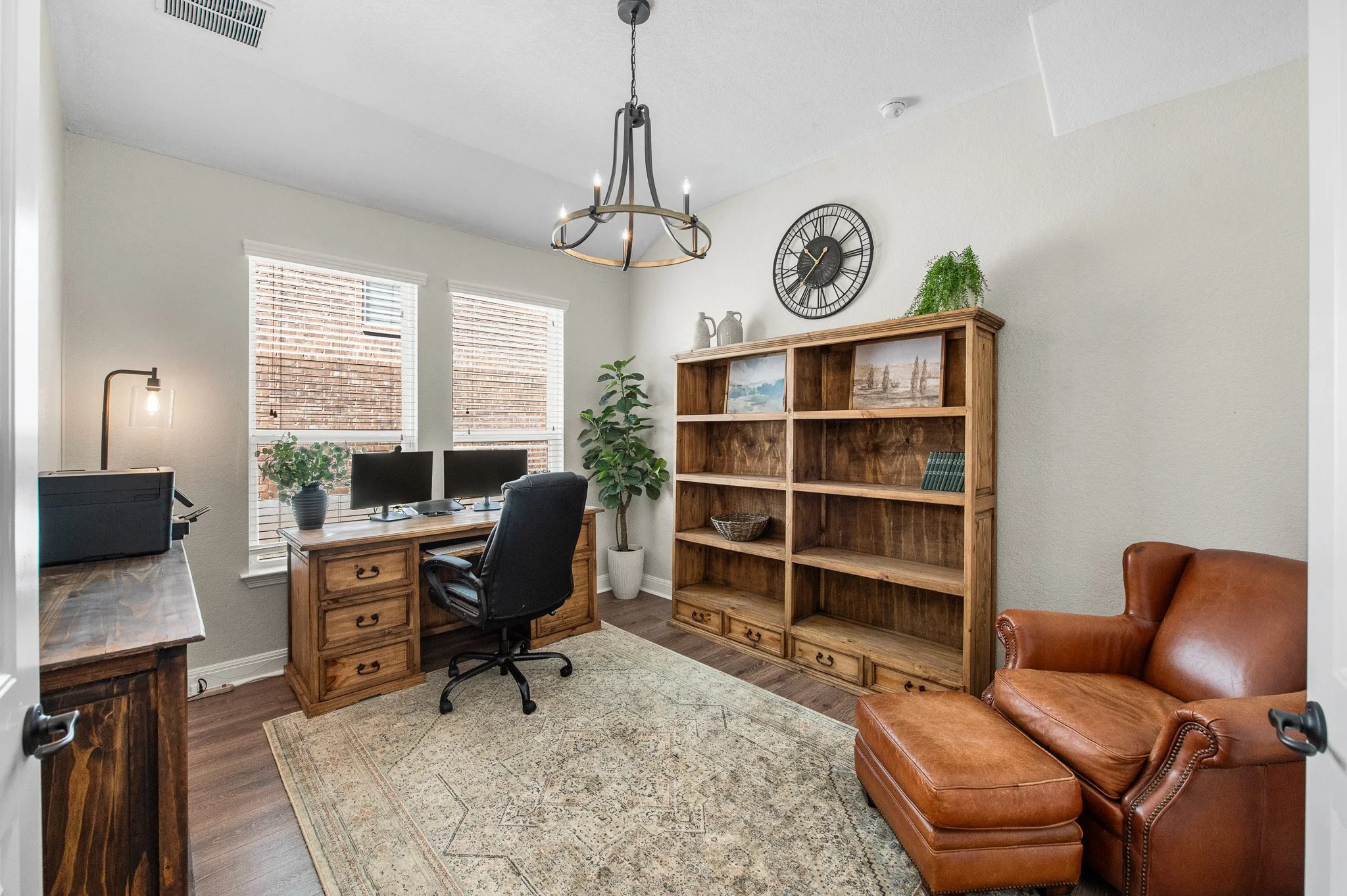 Home office with a wooden desk, dual computer monitors, black office chair, wooden bookshelves, leather armchair, rug, potted plants, and windows with blinds.