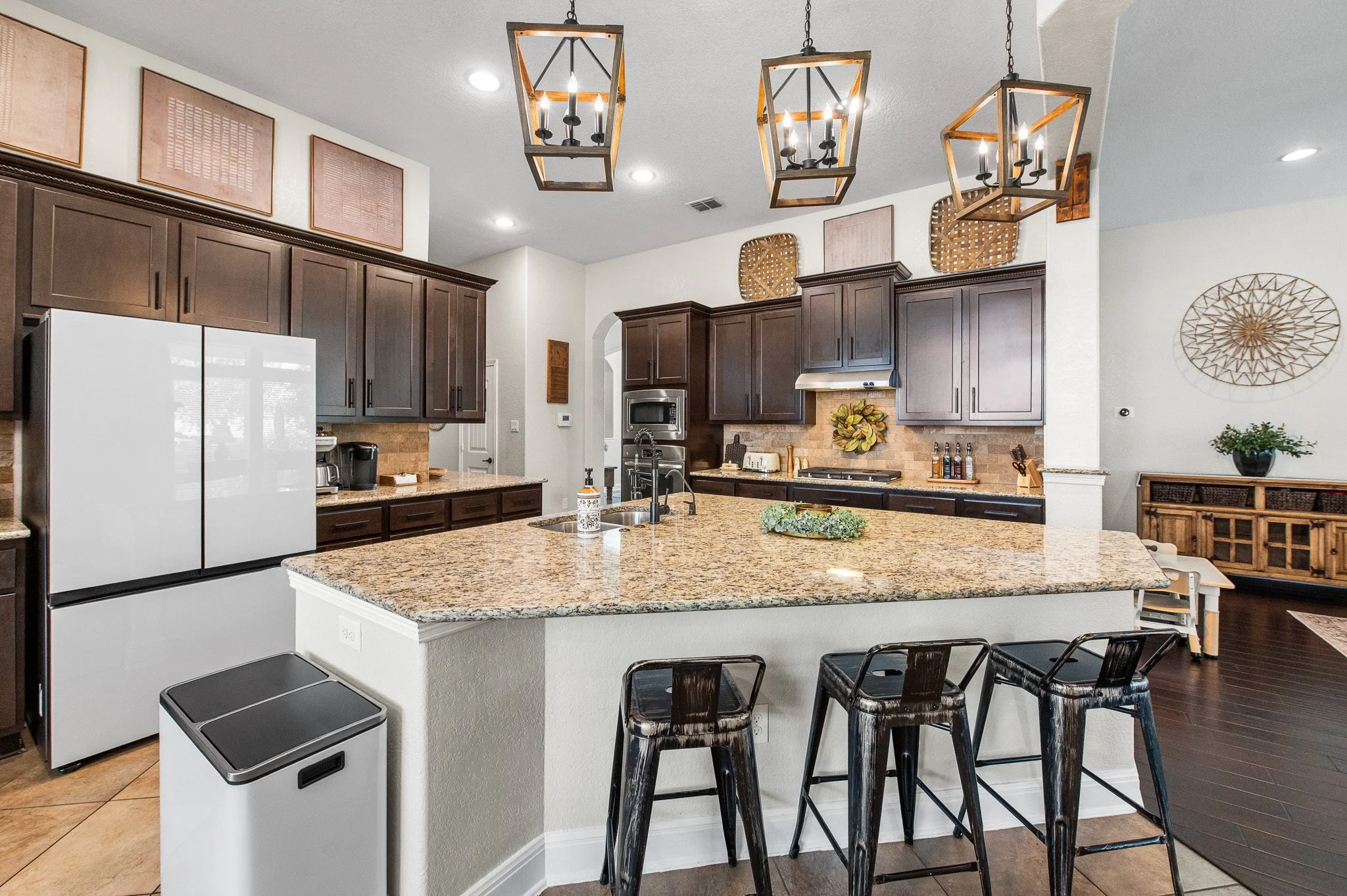 Modern kitchen with dark wood cabinets, granite countertops, a white refrigerator, and decorative lighting fixtures hanging above the island. There are three metallic barstools at the island and various kitchen appliances and decor on the countertops