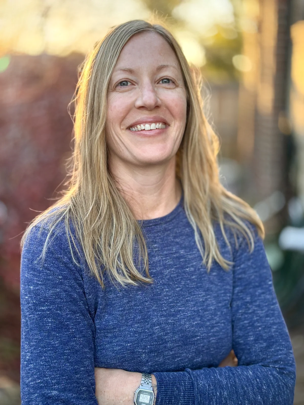 A woman with long blonde hair, blue eyes, and a big smile, standing outdoors in a park during sunset, wearing a blue long-sleeve shirt and a silver wristwatch.