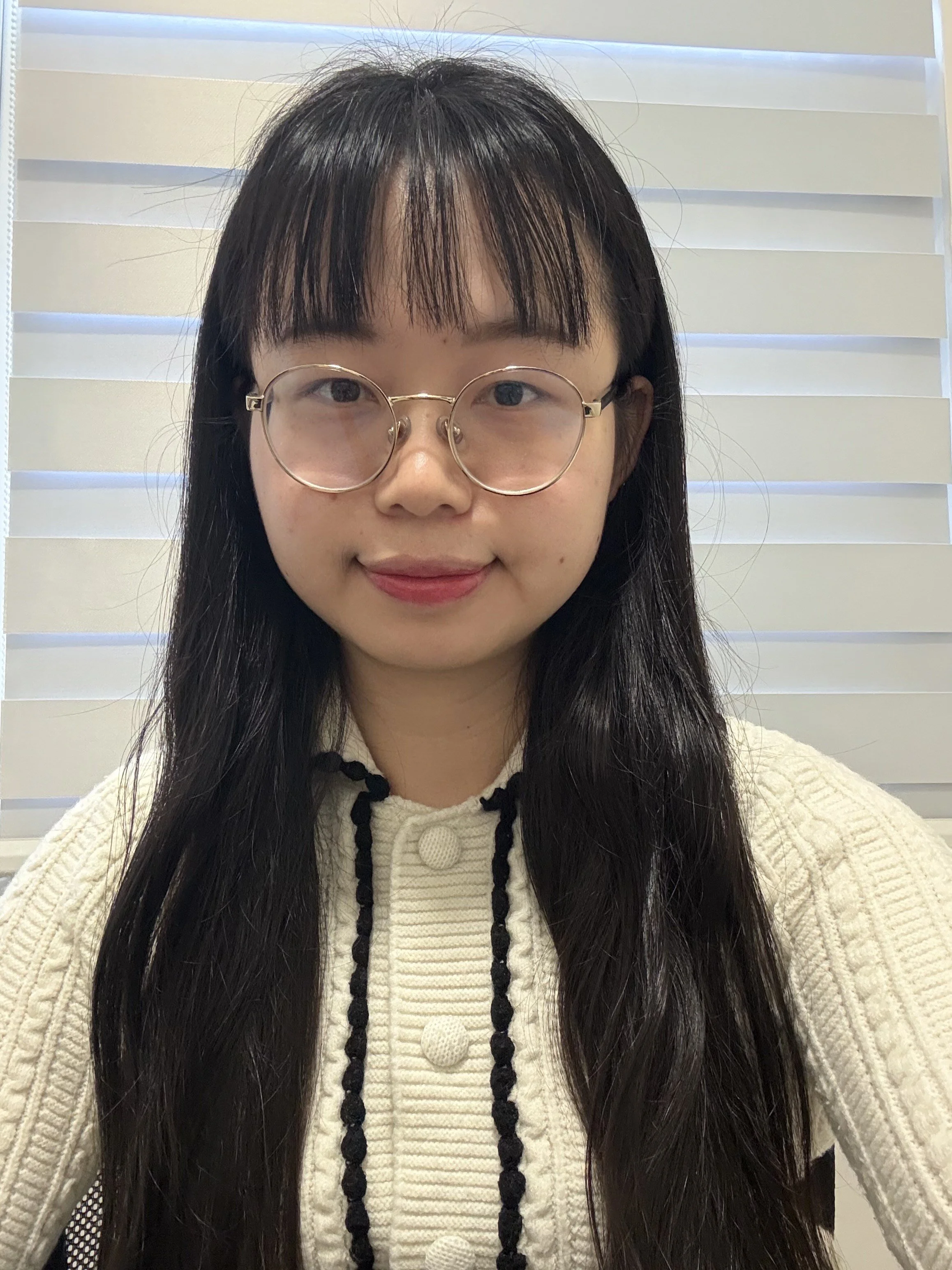 A young woman with long dark hair, wearing round glasses and a cream sweater with black detailing, sitting in front of white blinds.