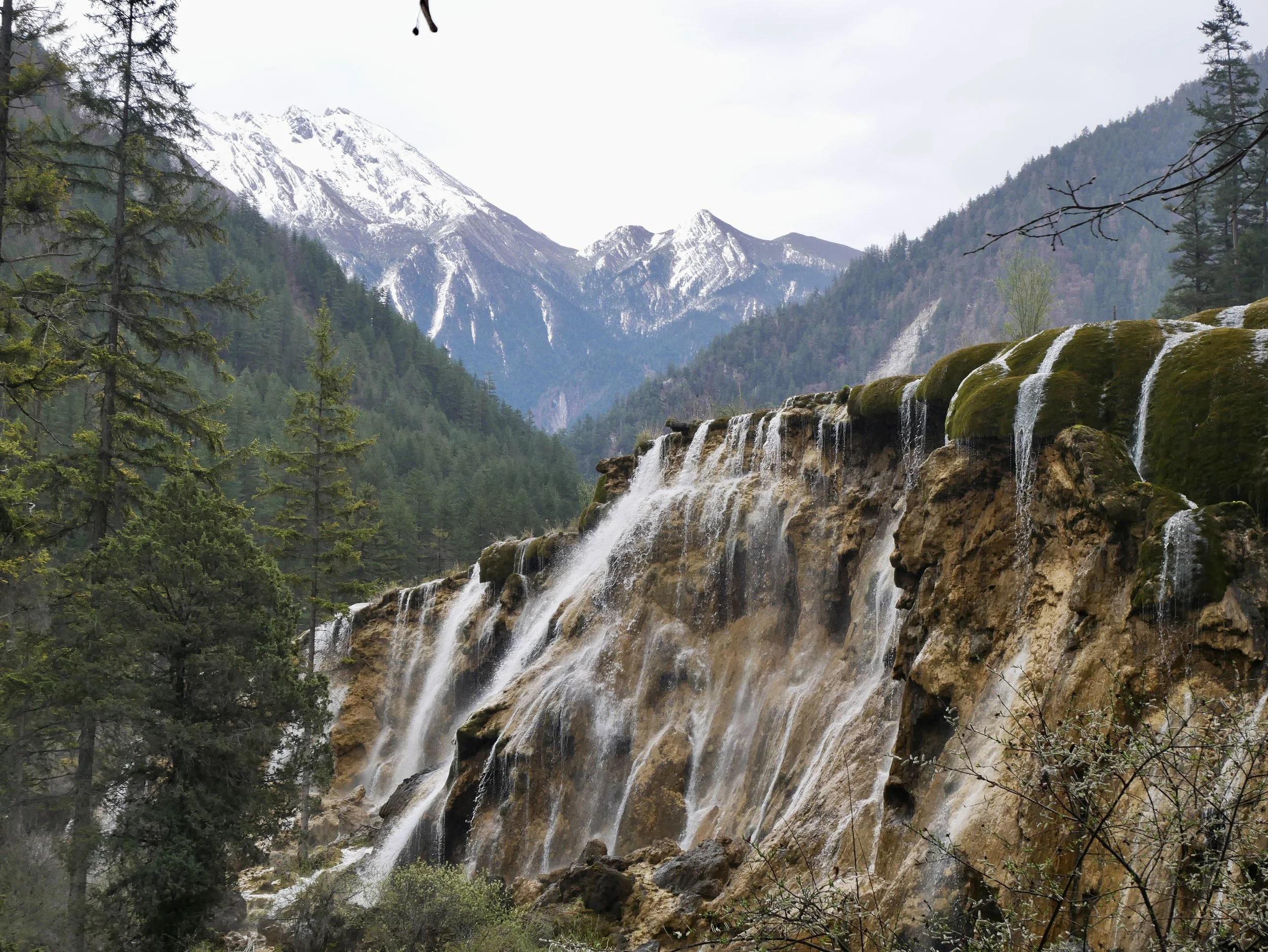 A scenic mountain landscape featuring snow-capped peaks, a lush forest, and a waterfall cascading over rocks in the foreground.