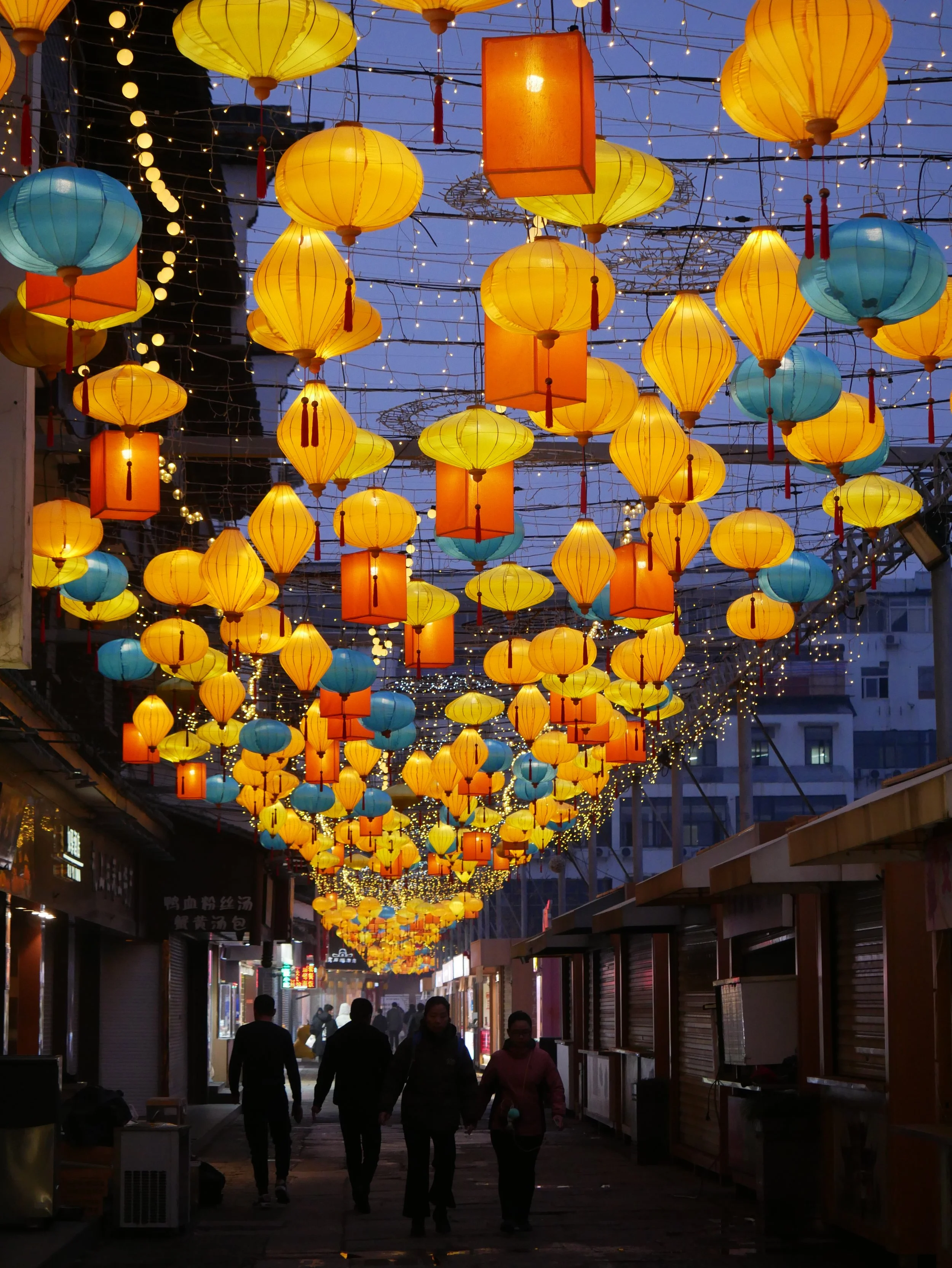 Street decorated with hanging yellow, orange, and blue lanterns and string lights, with people walking under the lanterns during evening or night.