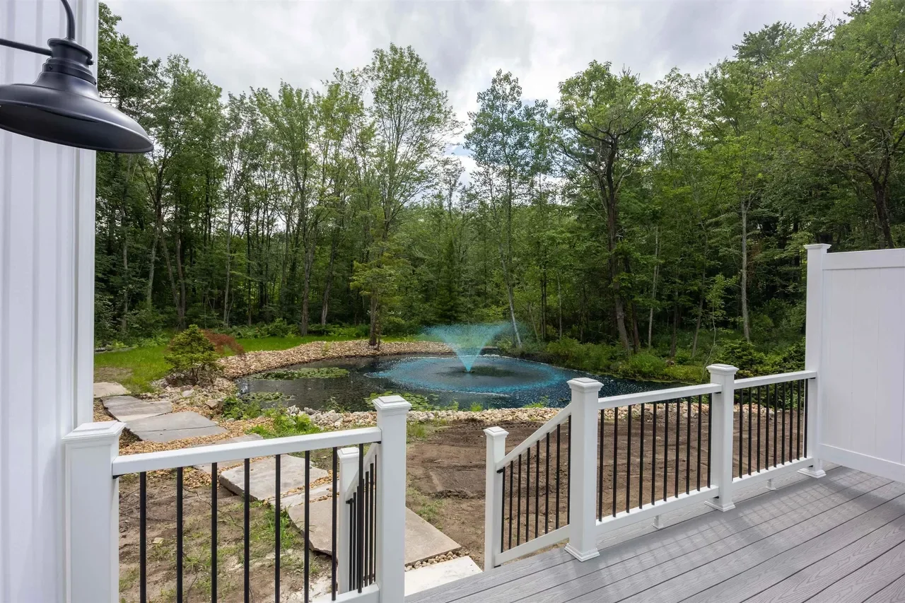 View from a deck overlooking a backyard pond with a fountain, surrounded by green trees and a wooded area.