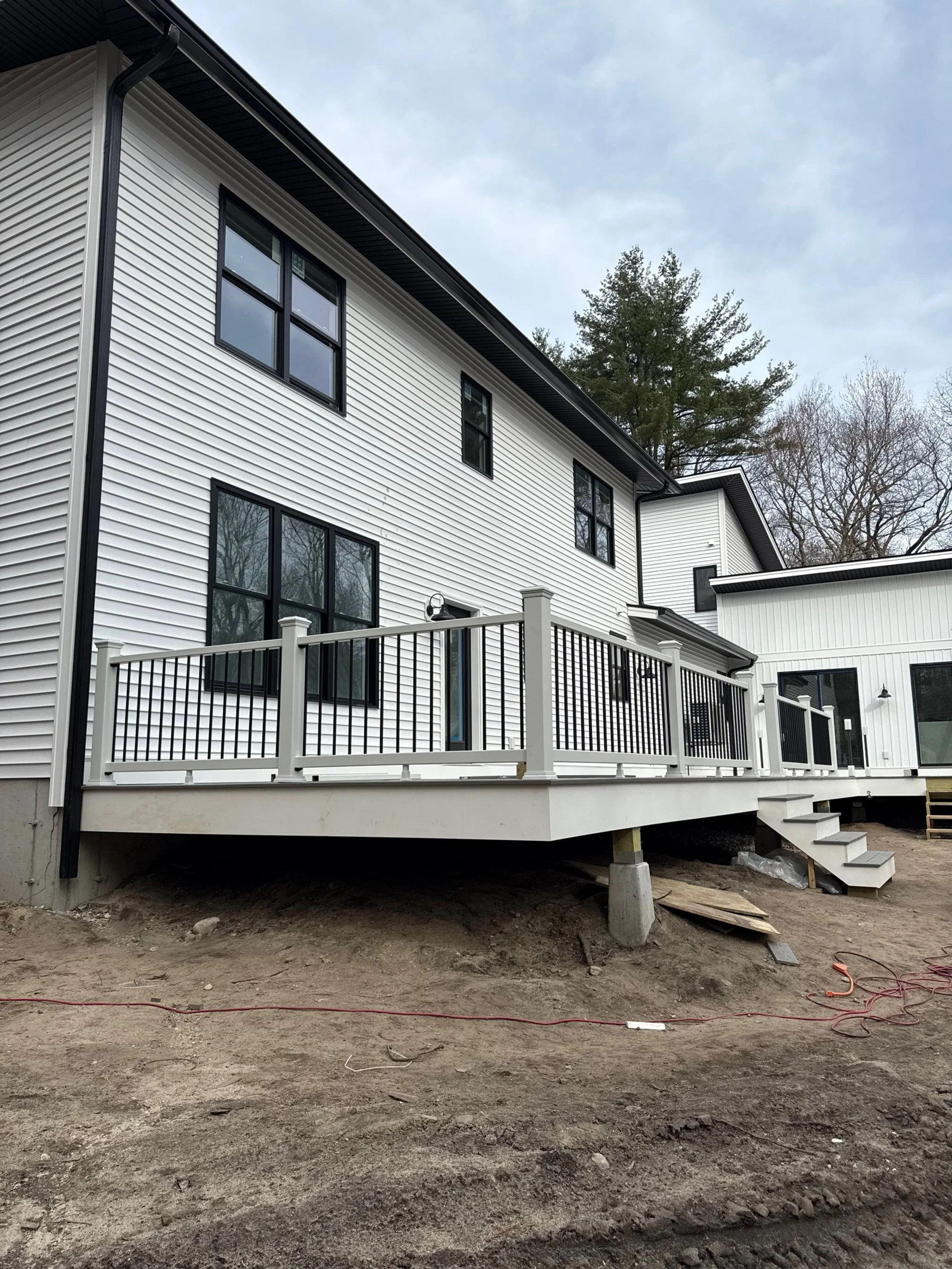 Newly built white house with black-trimmed windows and a wooden balcony, under construction, with exposed ground and construction materials underneath.