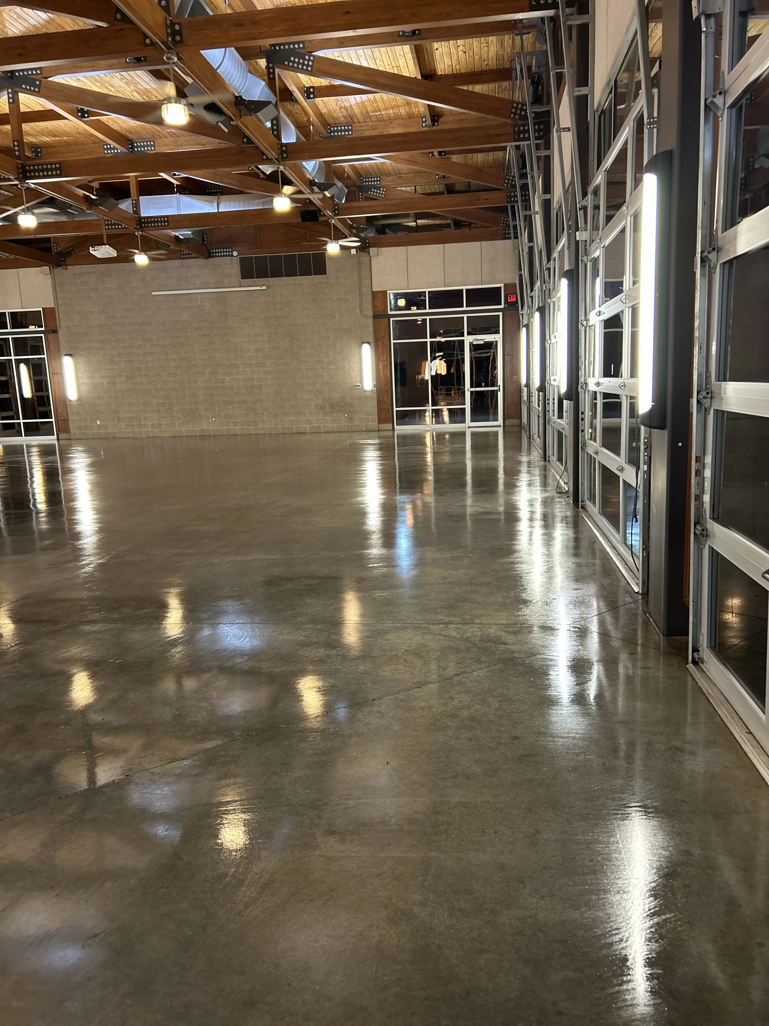 Empty industrial-style event space with a polished concrete floor, wooden ceiling beams, industrial lighting, and glass garage doors.