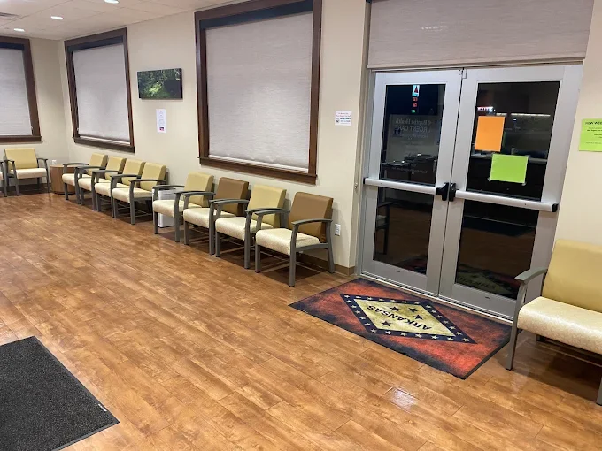 Waiting room with row of yellow and beige chairs along the wall, large windows with blinds, glass exit doors with signs, wooden flooring, and a decorative mat near the door.