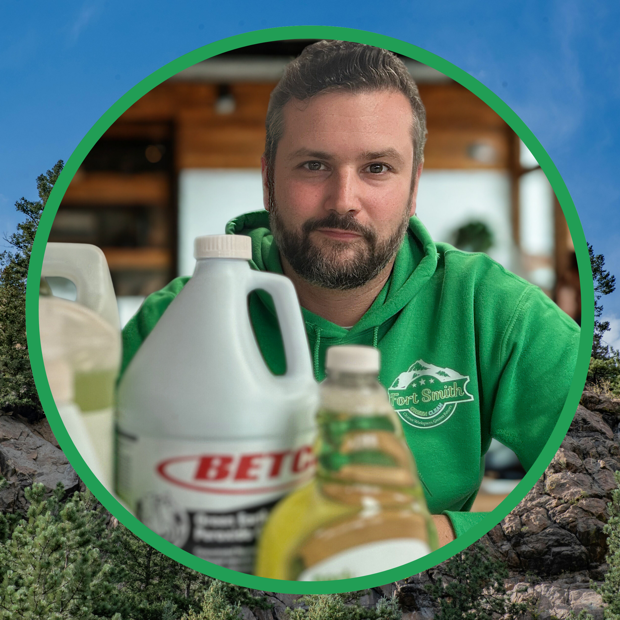 A man with a beard wearing a green hoodie sitting at a table with bottles of cleaning products, indoors with a blurred background