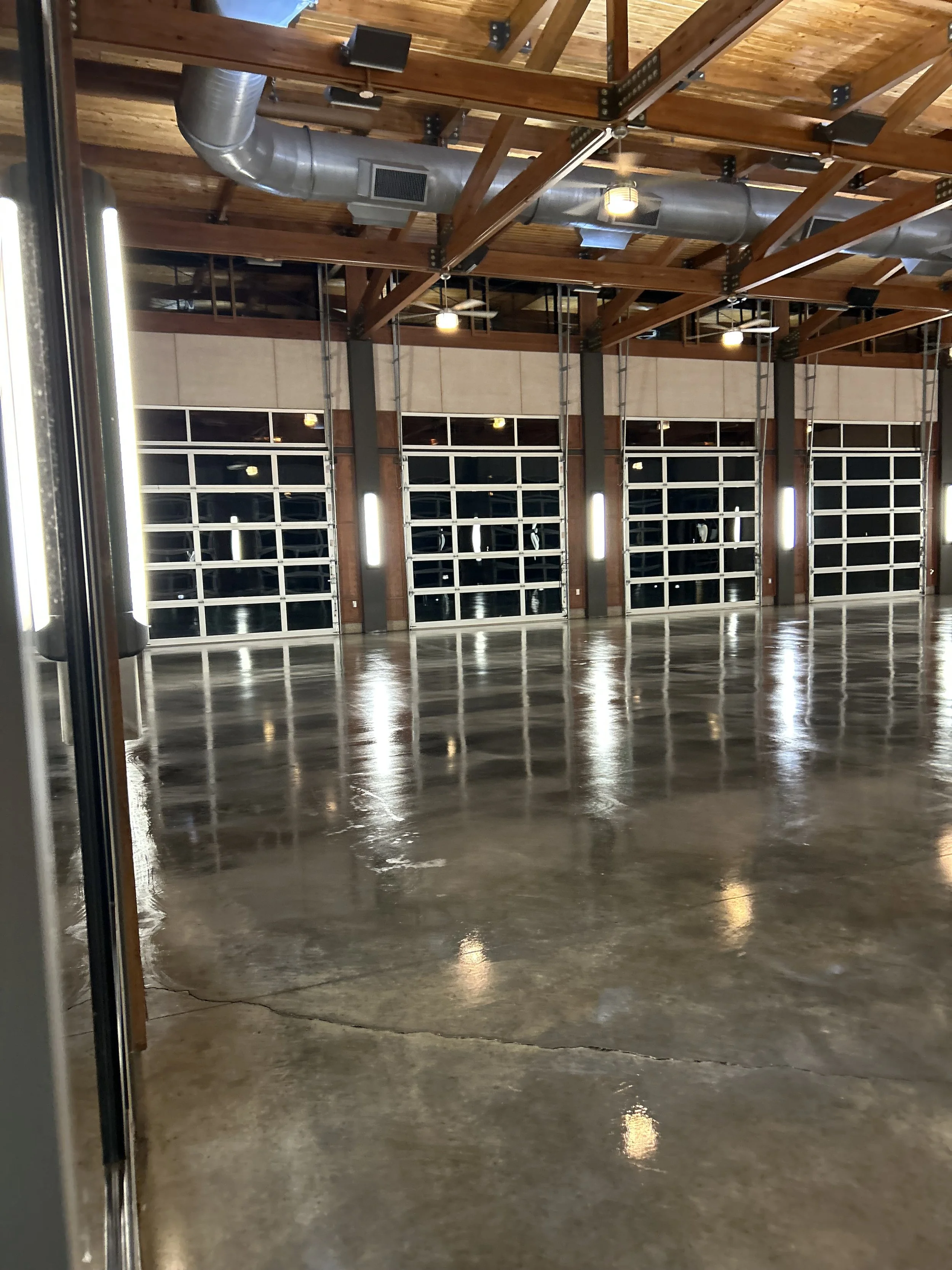 An empty industrial-style warehouse with polished concrete floors, large glass-paneled garage doors, exposed wooden beams and ceiling, and industrial ductwork.