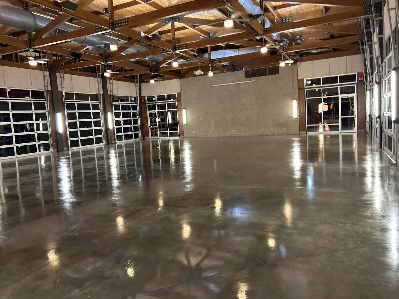 Empty indoor space with polished concrete floor, large glass garage-style doors, wooden ceiling beams, and industrial lighting.