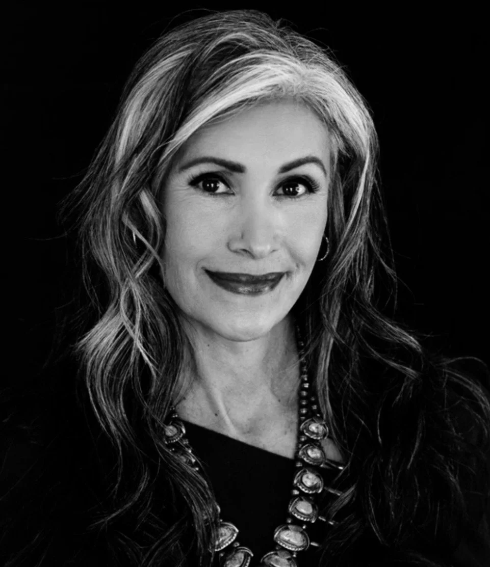 Black and white portrait of a woman with long, wavy hair and a friendly expression, wearing a necklace with large gemstones.