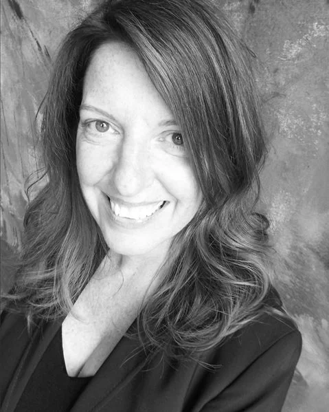 A woman with wavy, shoulder-length hair smiling in front of a textured wall.
