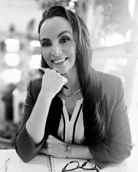 A woman with long hair smiling, resting her chin on her hand, sitting at a table with glasses and a notebook.