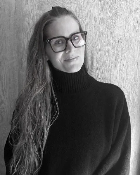 A woman with long hair wearing glasses and a black turtleneck sweater, standing against a wooden background.