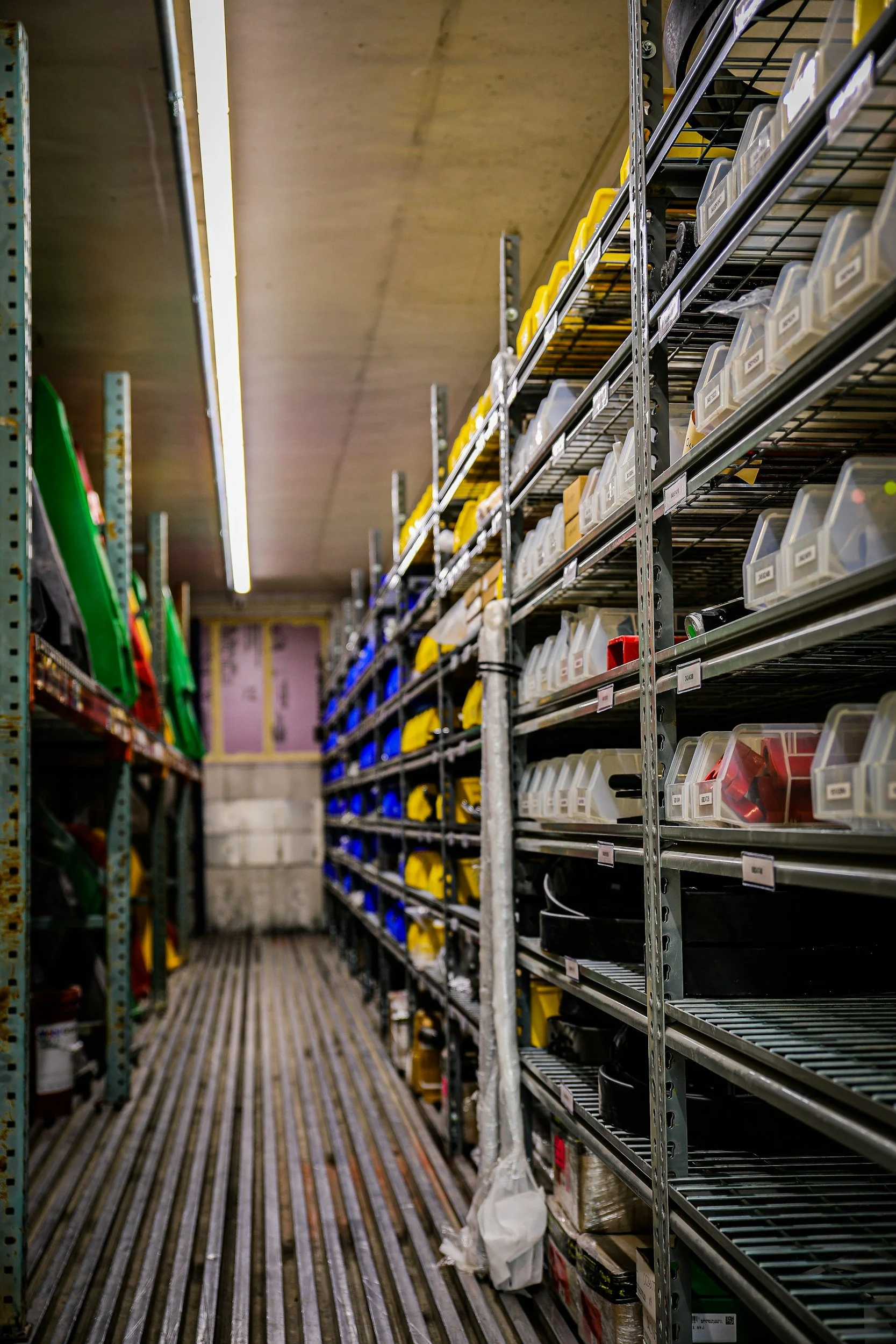Industrial storage room with metal shelving filled with plastic bins and containers, some labeled, along with large plastic storage sleds on the left.