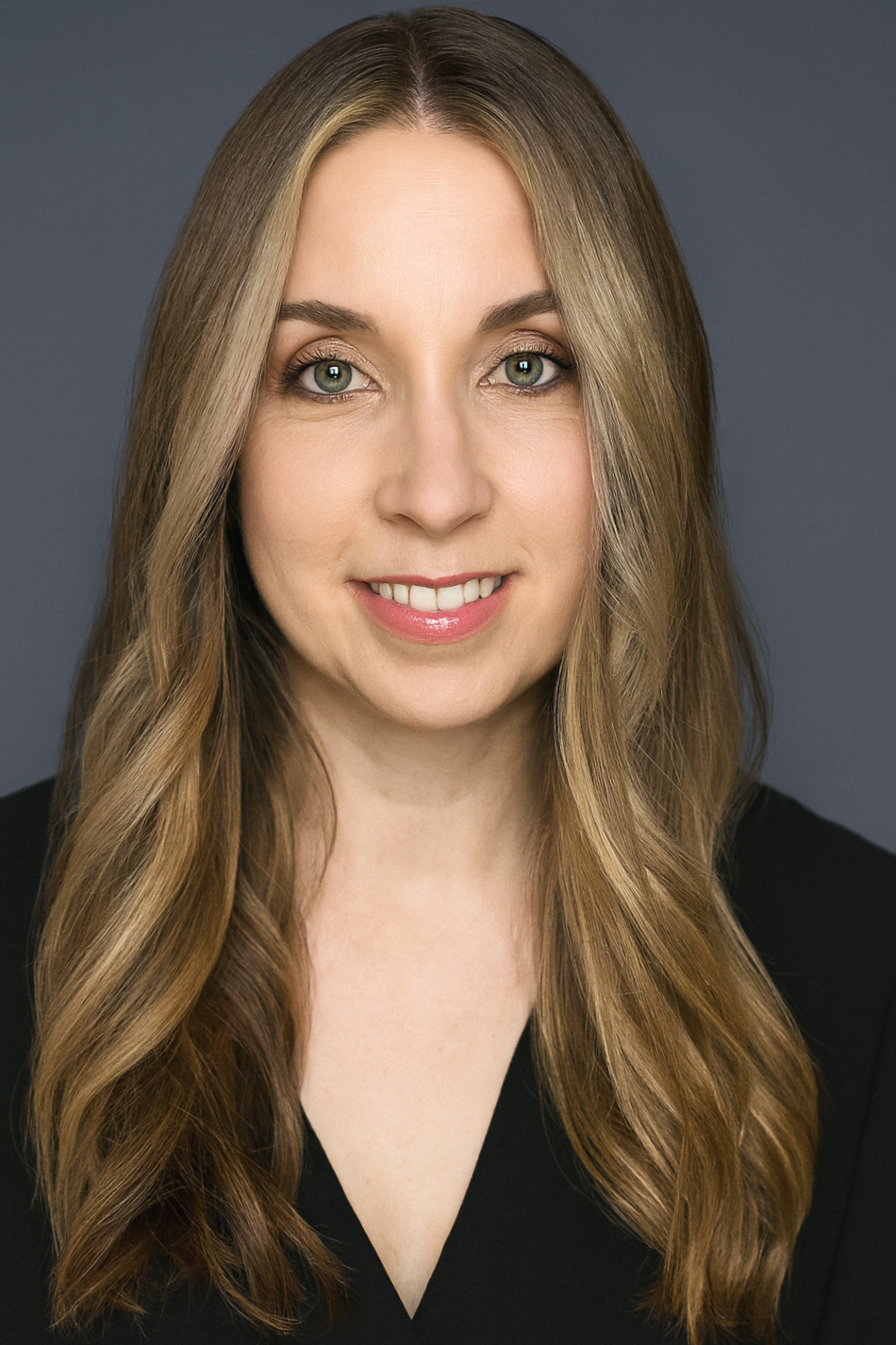 A woman with long, wavy blonde hair and green eyes, smiling, wearing a black top with a V-neck, in front of a gray background.