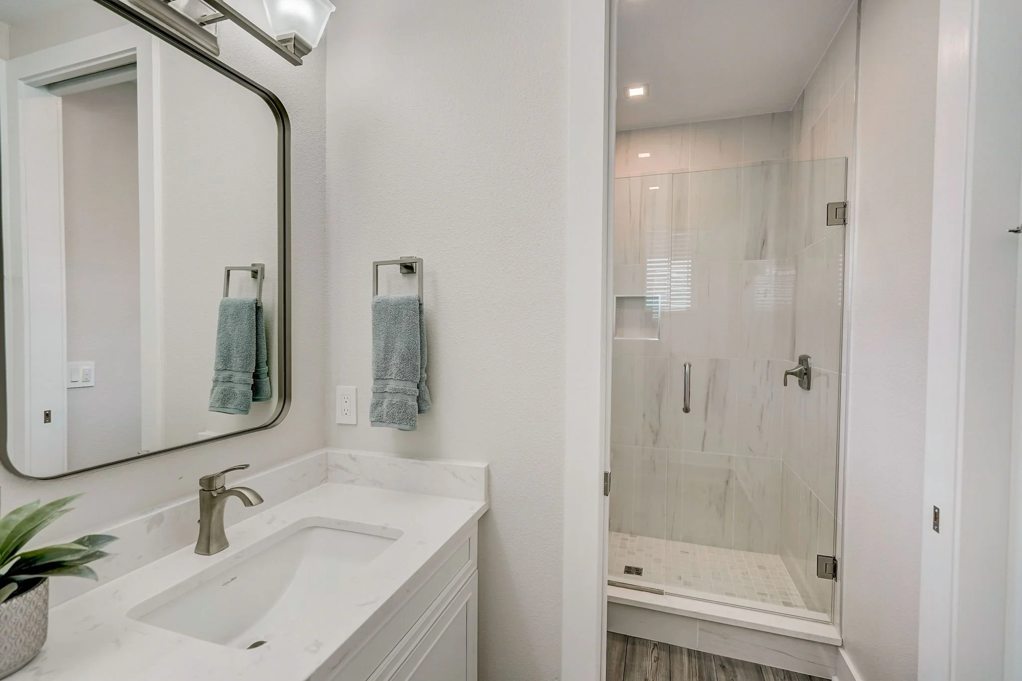 Modern bathroom with a white vanity, framed mirror, gray towels, and a glass-enclosed shower with marble tile walls.