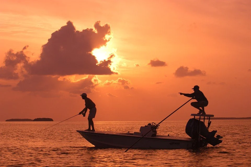 Silhouettes of two people fishing from a boat at sunset with a colorful sky and calm water.