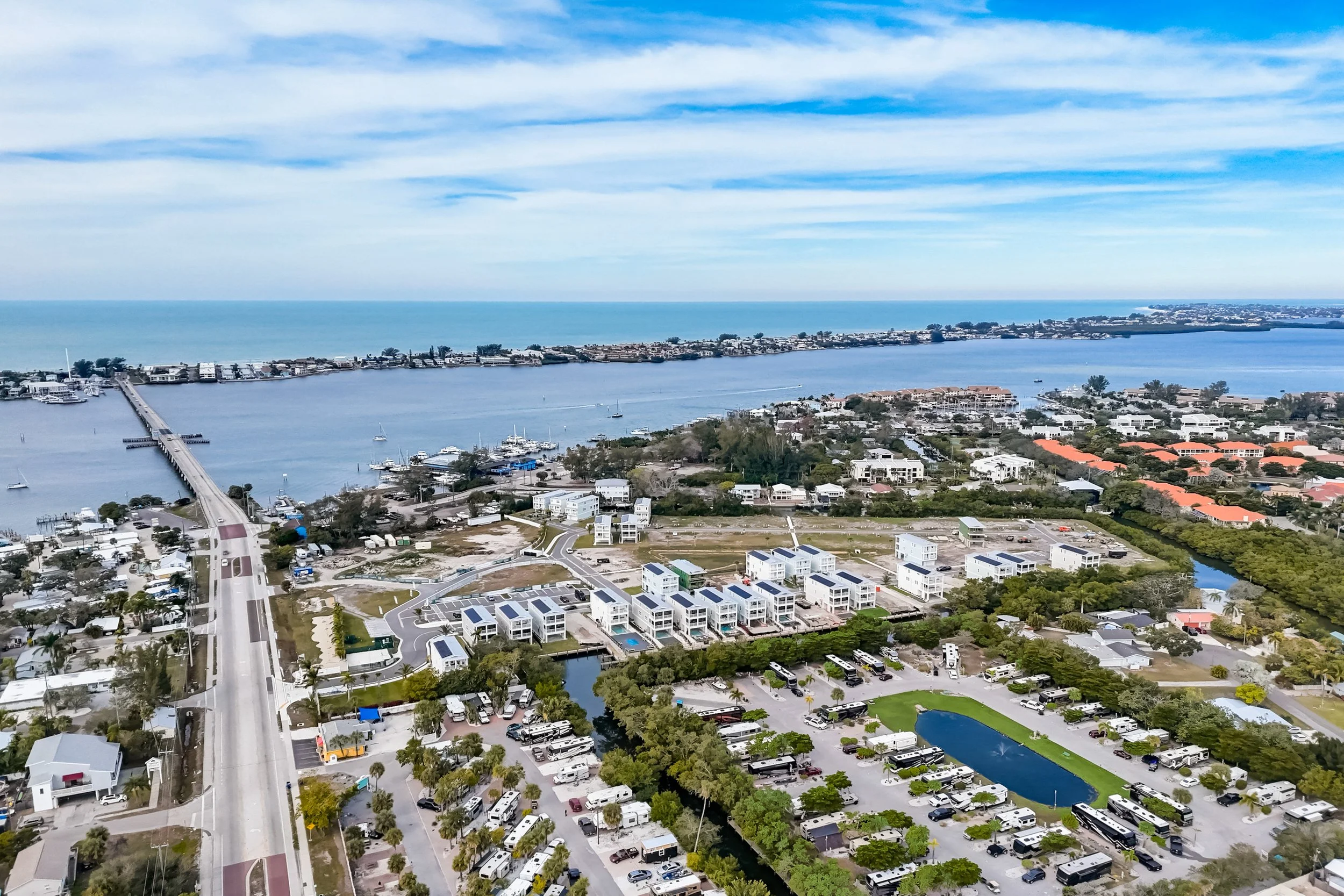 Aerial view of a coastal city with a bridge over a waterway, marina filled with boats, modern residential buildings with solar panels, a parking lot, a small pond, and the ocean in the background under a partly cloudy sky.