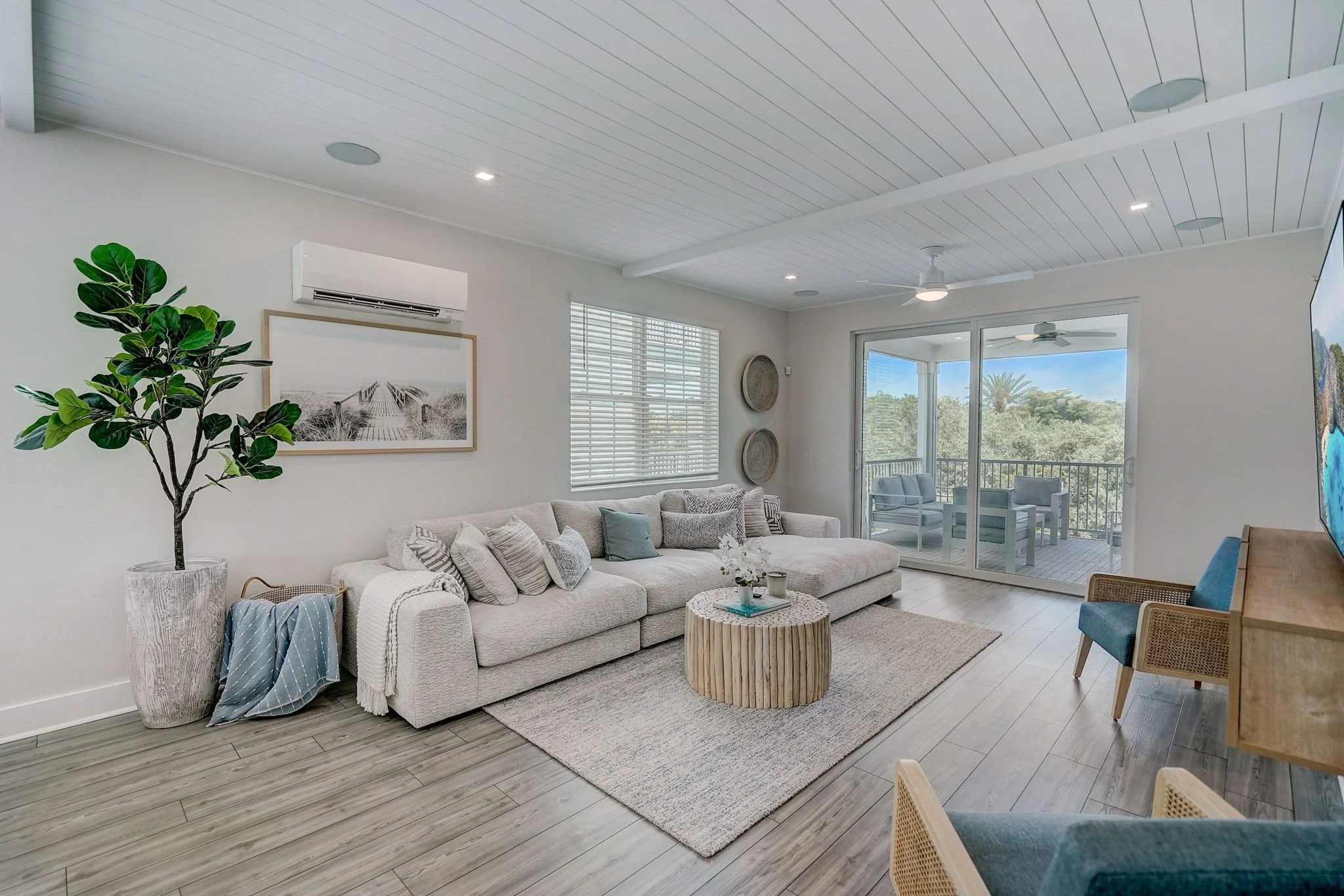 Bright living room with a large beige sofa, a wooden coffee table, and a sliding glass door leading to a balcony with outdoor seating and a view of trees. Decor includes a large potted plant, framed artwork, and woven wall accents.