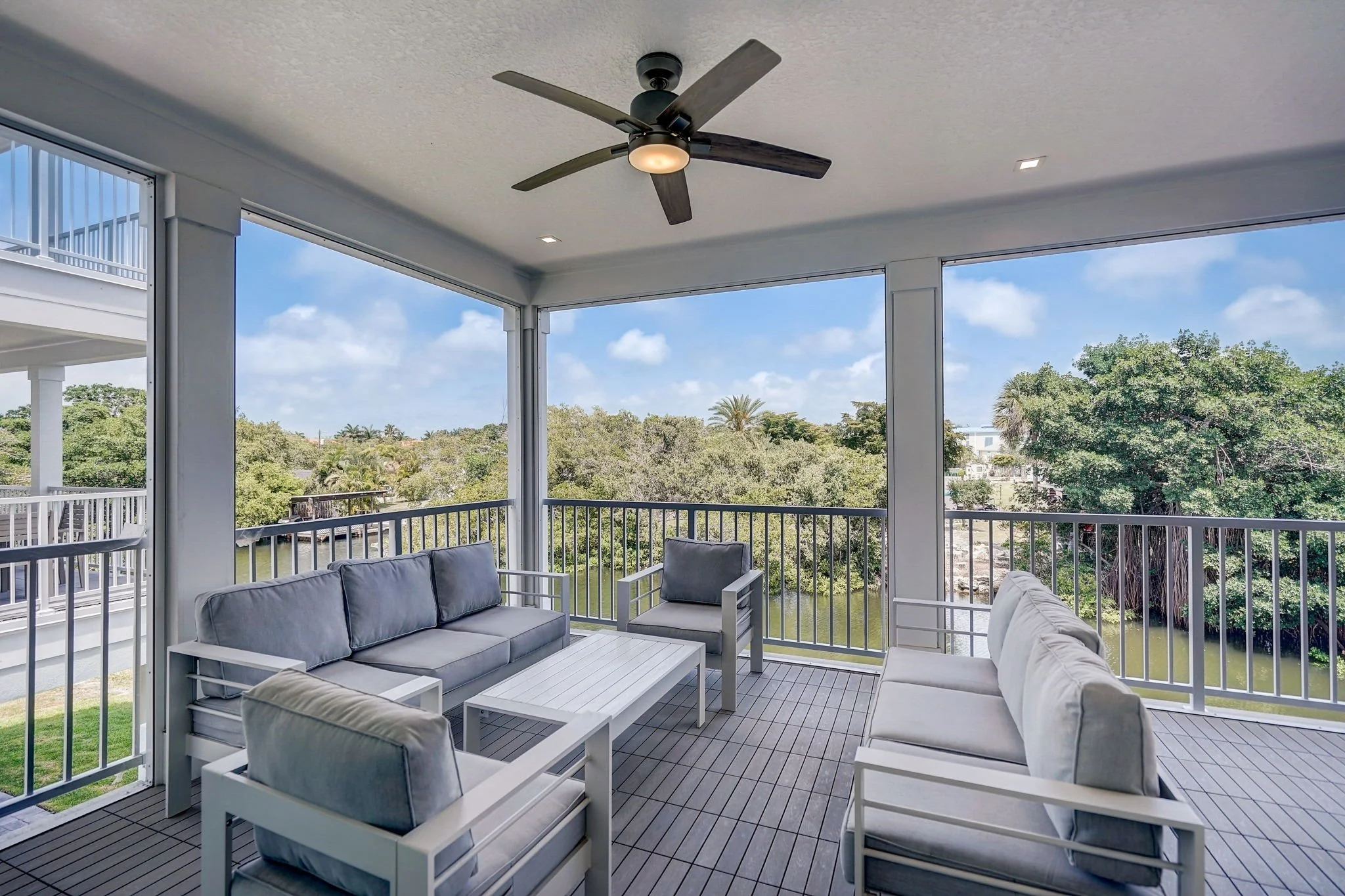 Outdoor porch with white furniture, including sofas and chairs, surrounded by a white railing, overlooking a canal with lush trees and a blue sky with scattered clouds.