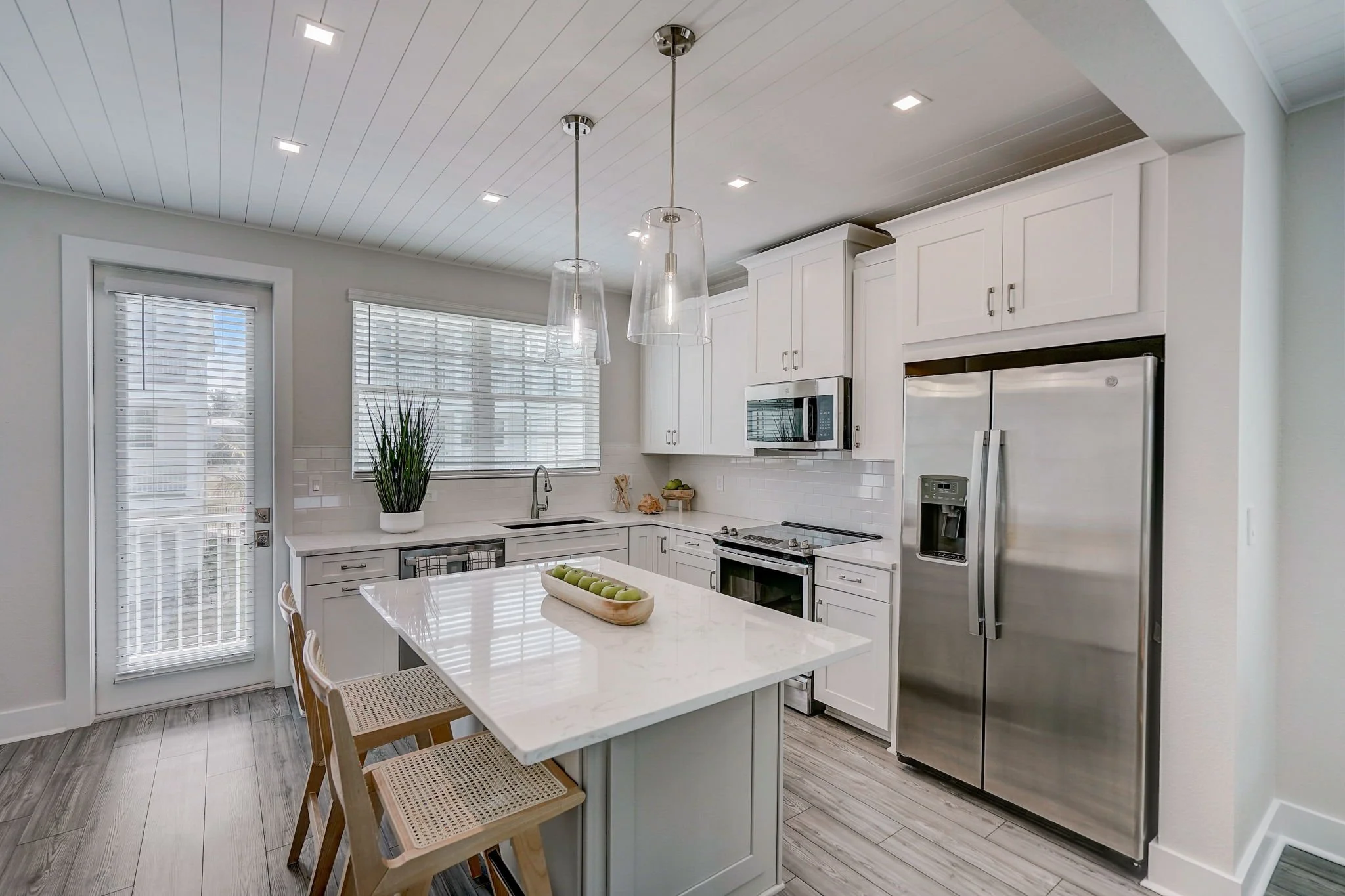 Modern white kitchen with island, stainless steel refrigerator, microwave, and oven, large window with blinds, door leading outside, light wood flooring, and pendant lights over the island.