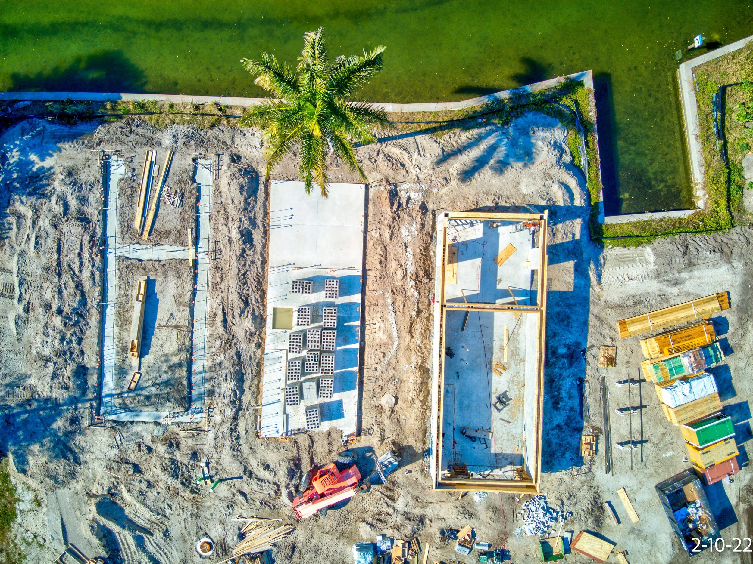 Aerial view of a construction site next to a body of water, with building foundations, construction materials, and a palm tree.