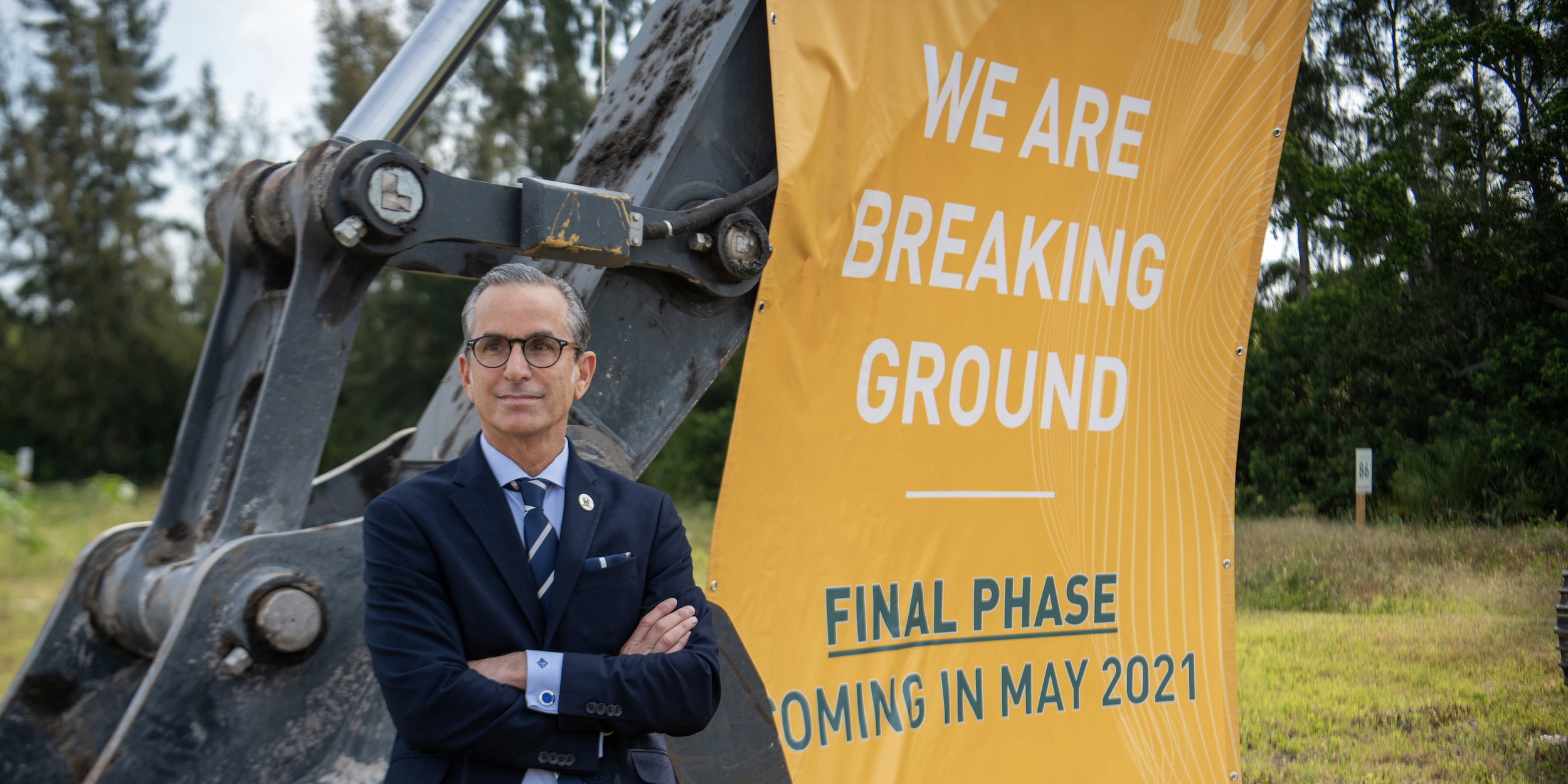 A man in a suit and glasses standing with arms crossed near a construction excavator and a yellow sign indicating that a project is in its final phase