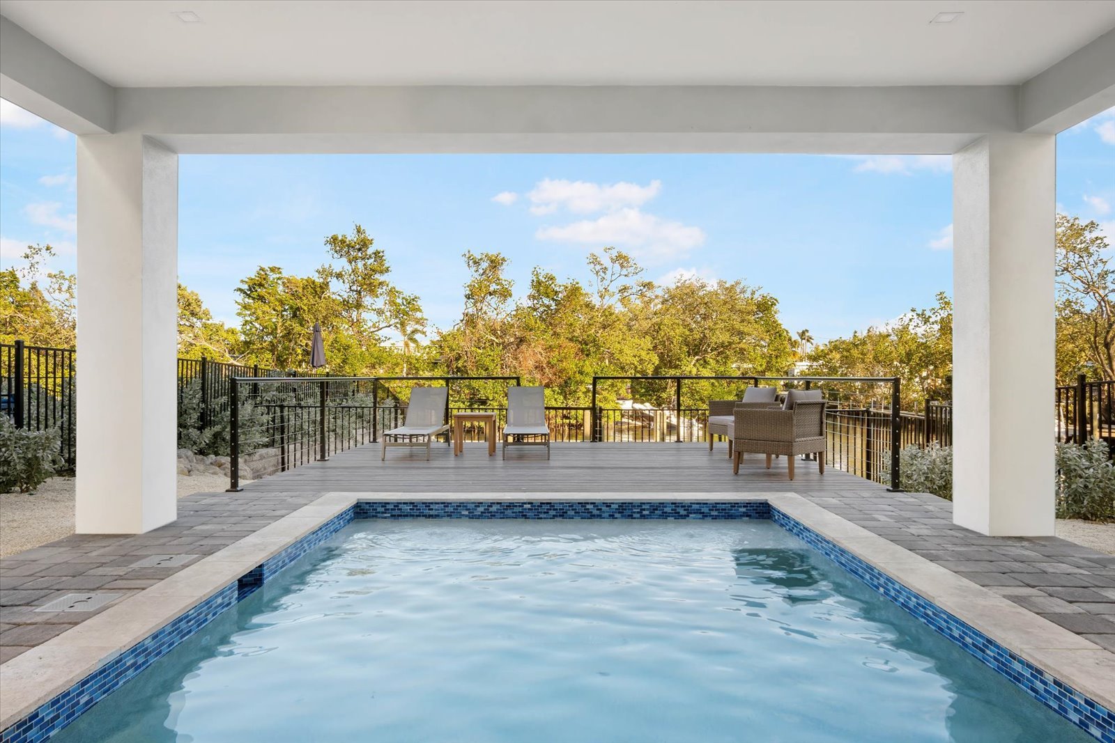 View from a covered patio showing a backyard swimming pool with lounge chairs, a small table, and outdoor seating, surrounded by a black metal fence, with trees and a blue sky in the background.