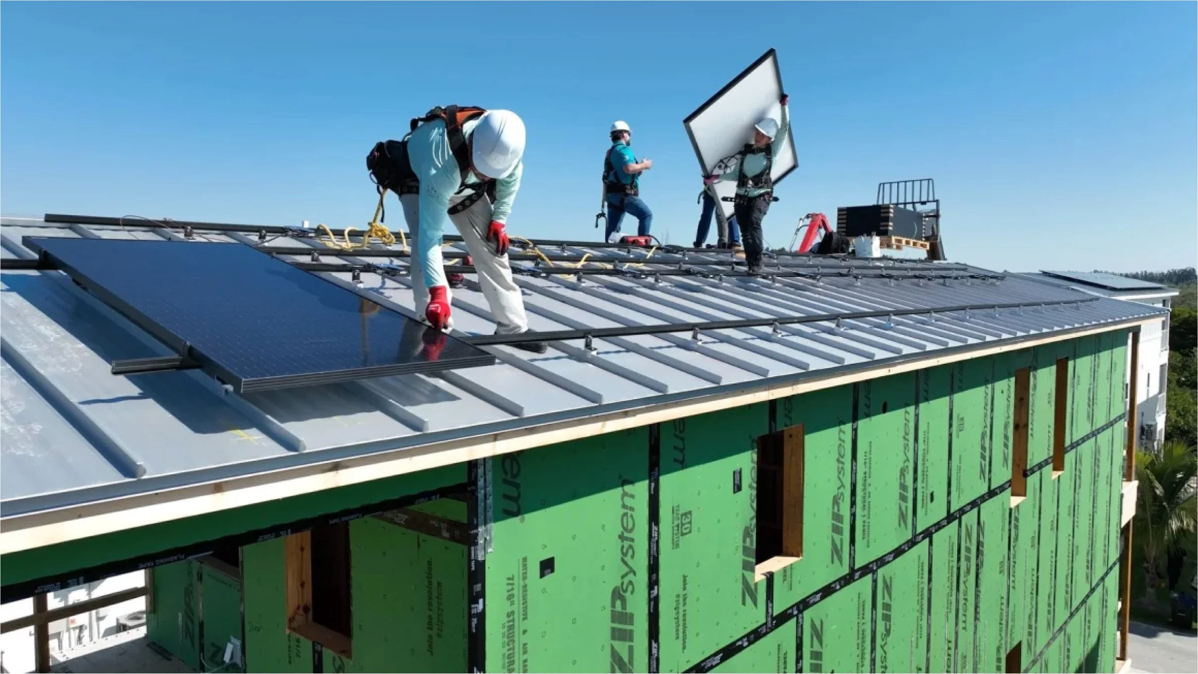 Construction workers installing solar panels on the roof of a building under clear blue sky, with green sheathing on the exterior walls.