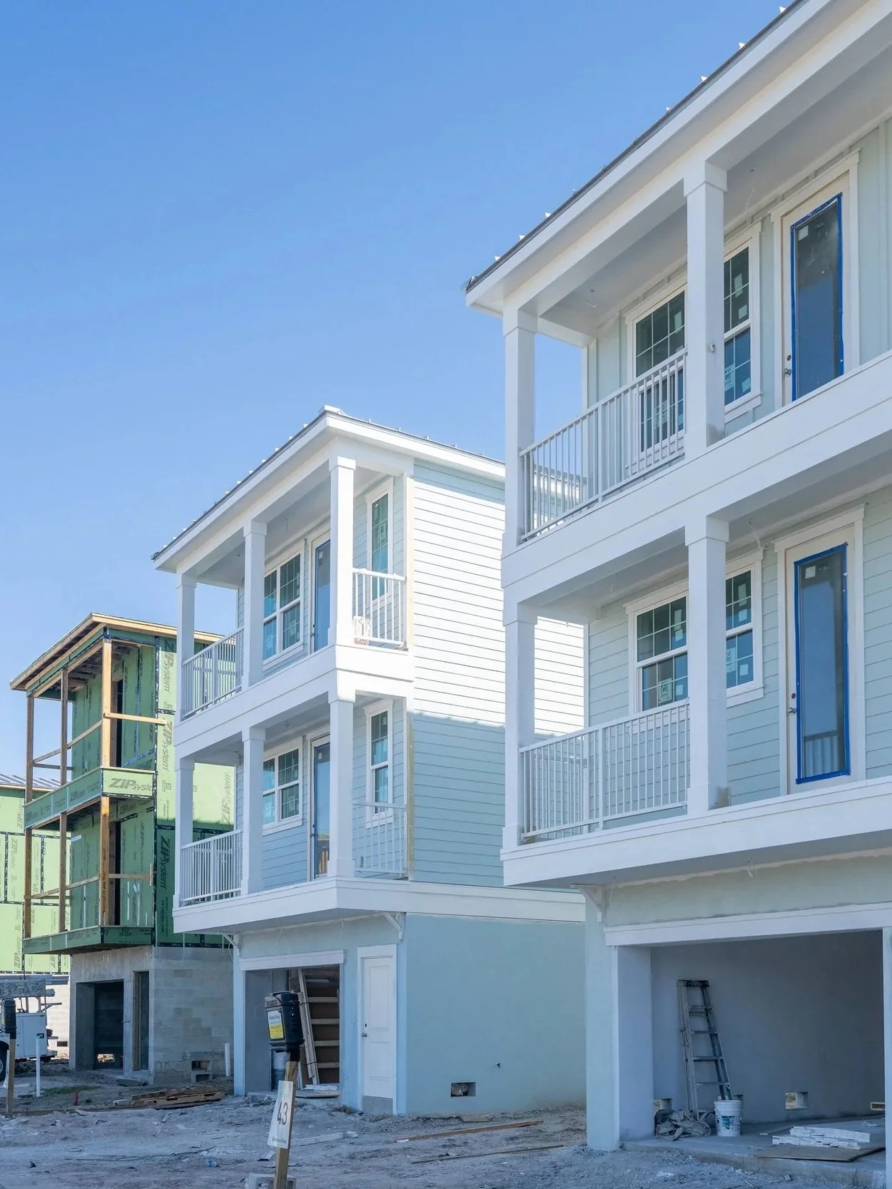 Under construction multi-story residential building with white siding and small balconies, next to a smaller house under construction with green sheathing, construction materials on the ground, and a ladder leaning against the building.