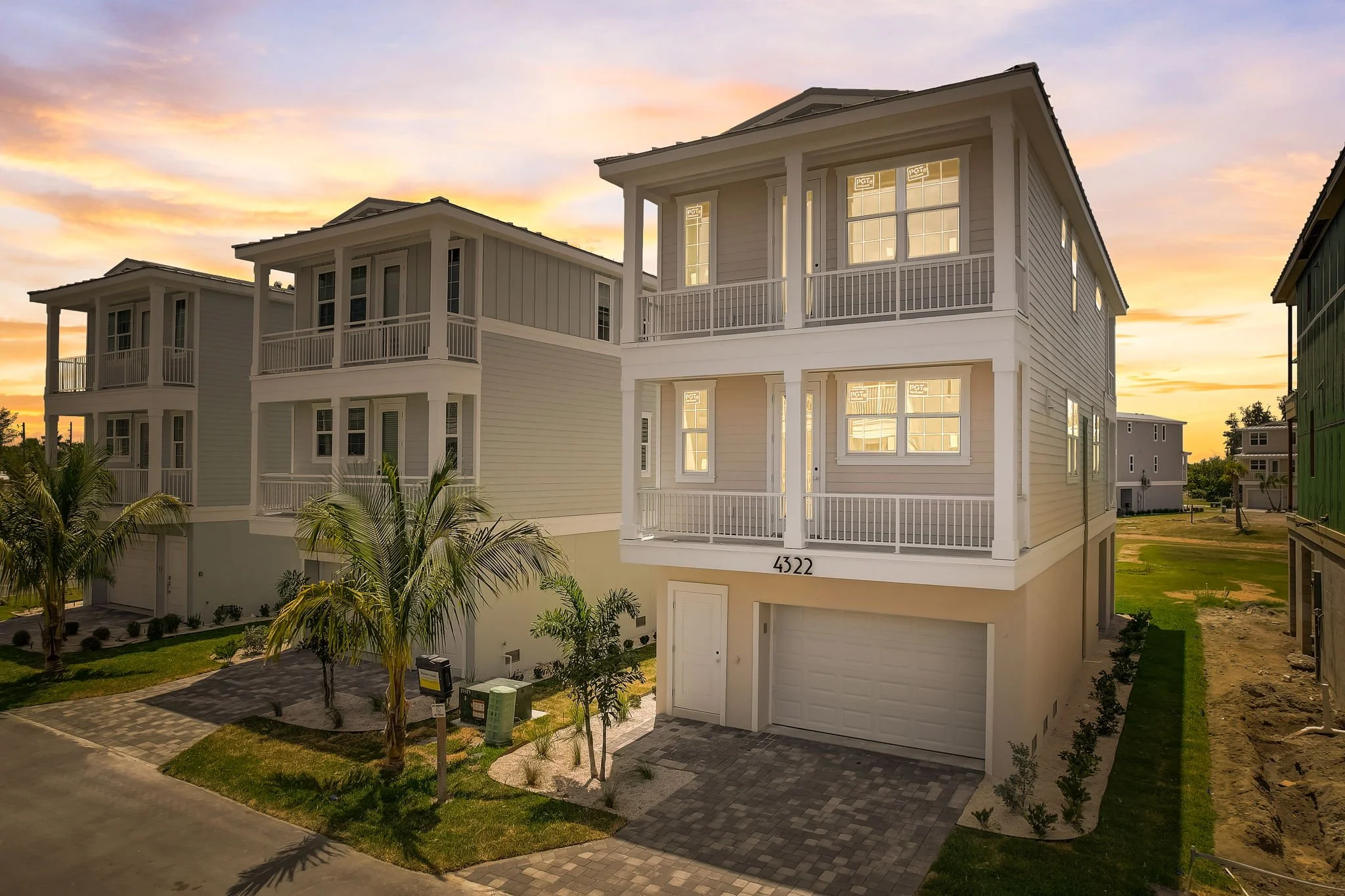 A modern multi-story white house with a garage, porch, and balconies, surrounded by a landscaped yard and palm trees, during sunset in a residential neighborhood.