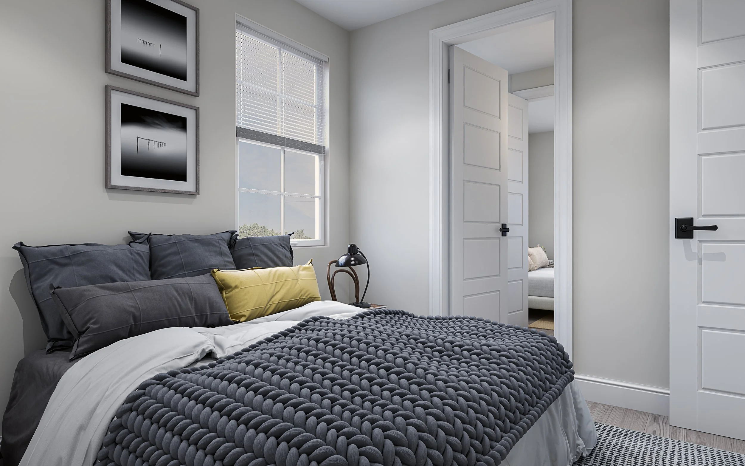 A bedroom with a bed covered in dark gray bedding and multiple pillows, including a yellow one, beside a nightstand with a black lamp. There are two framed black and white pictures on the wall, a window with white blinds, and a door leading to anothe