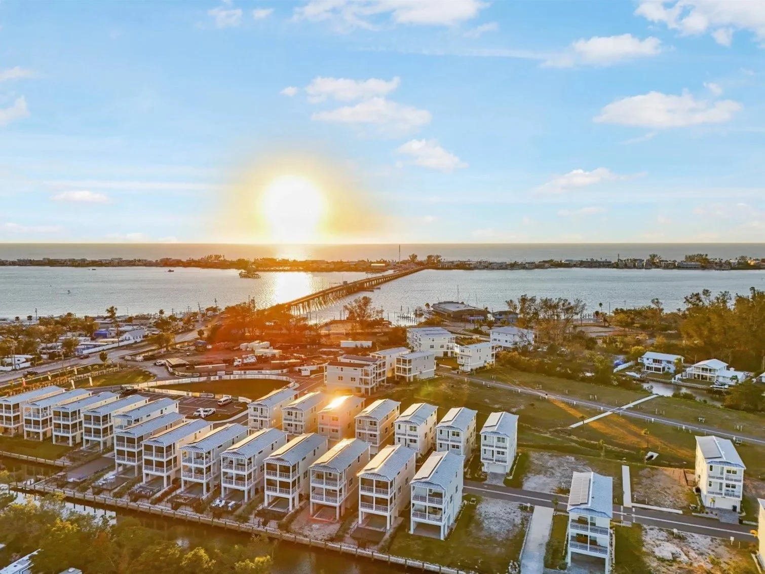 Aerial view of a coastal residential area during sunset, with white houses, a bridge over water, and the ocean in the background.