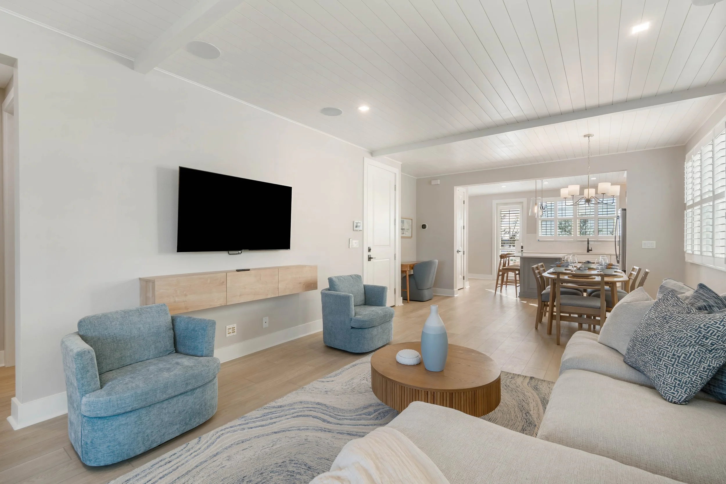 Living room with a wall-mounted TV, gray chairs, a beige sofa, a wooden coffee table with vases, and a dining area with a round table and chairs, bright natural light, and white walls.