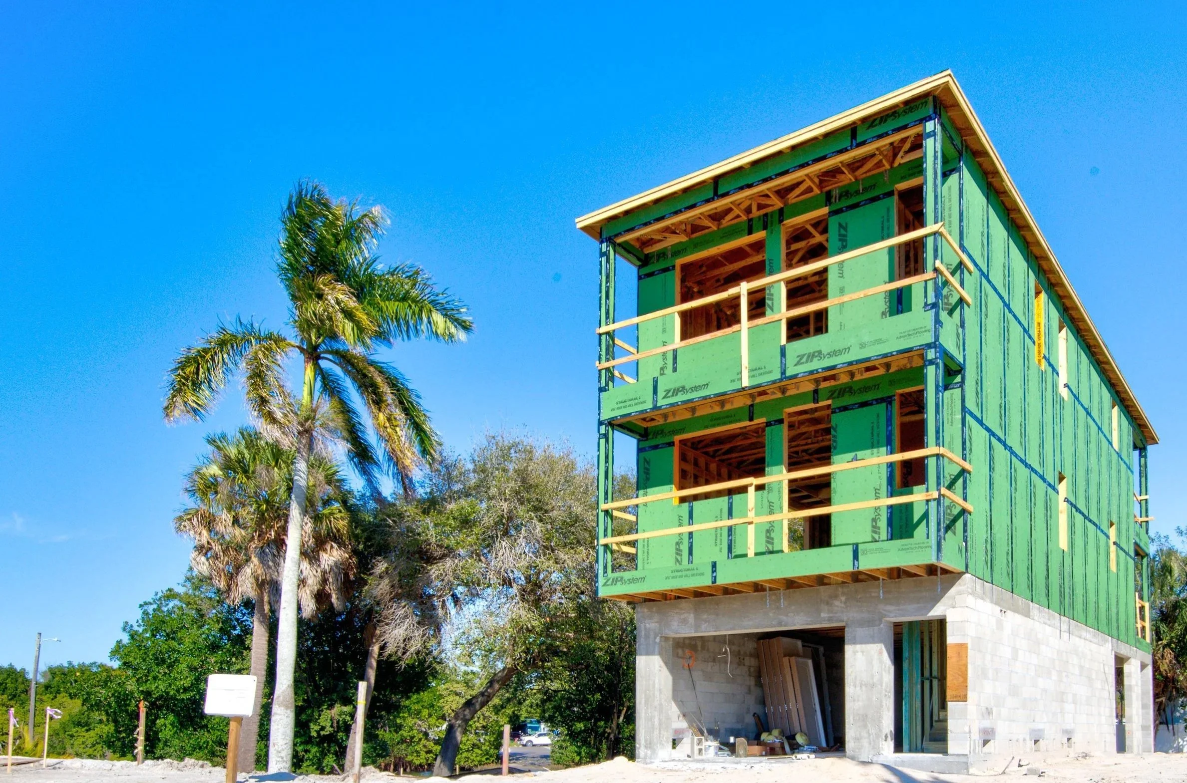 A three-story house under construction with a concrete lower level and green sheathing on the upper floors, surrounded by tropical trees and a clear blue sky.