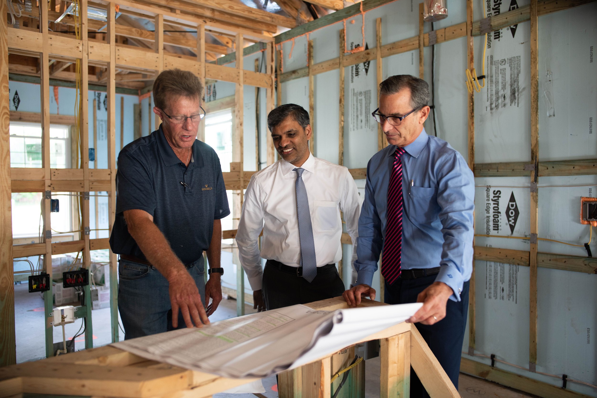 Three men examining blueprints inside a house under construction, with exposed wooden framing and insulation.