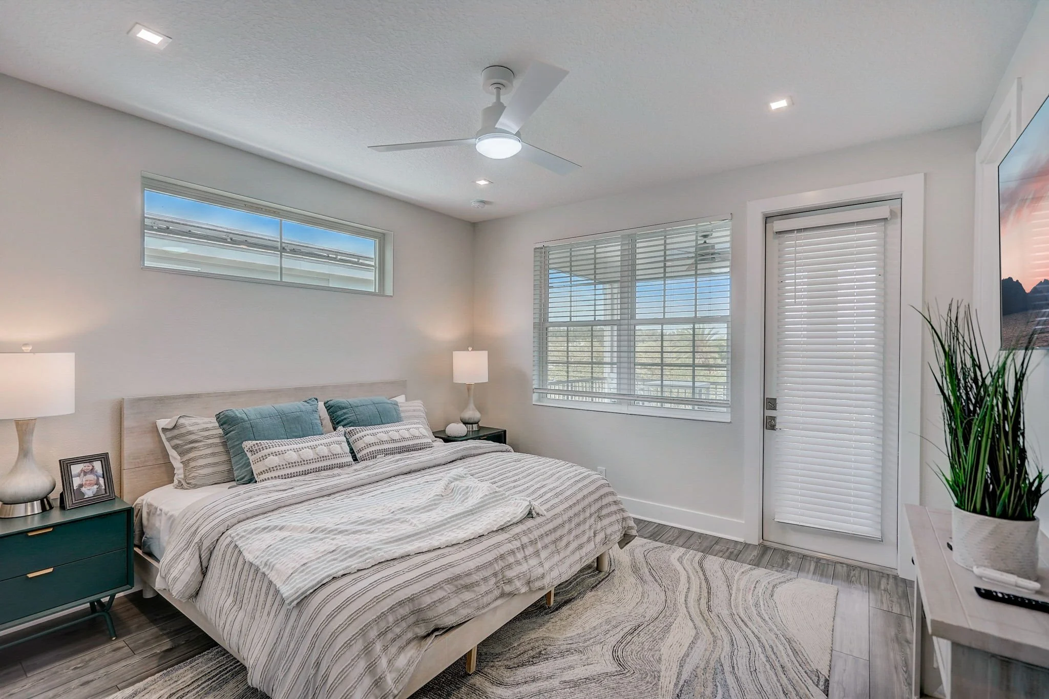 A cozy bedroom with a large bed, blue pillows, side tables with lamps, a window with blinds, a door with blinds, and a ceiling fan.