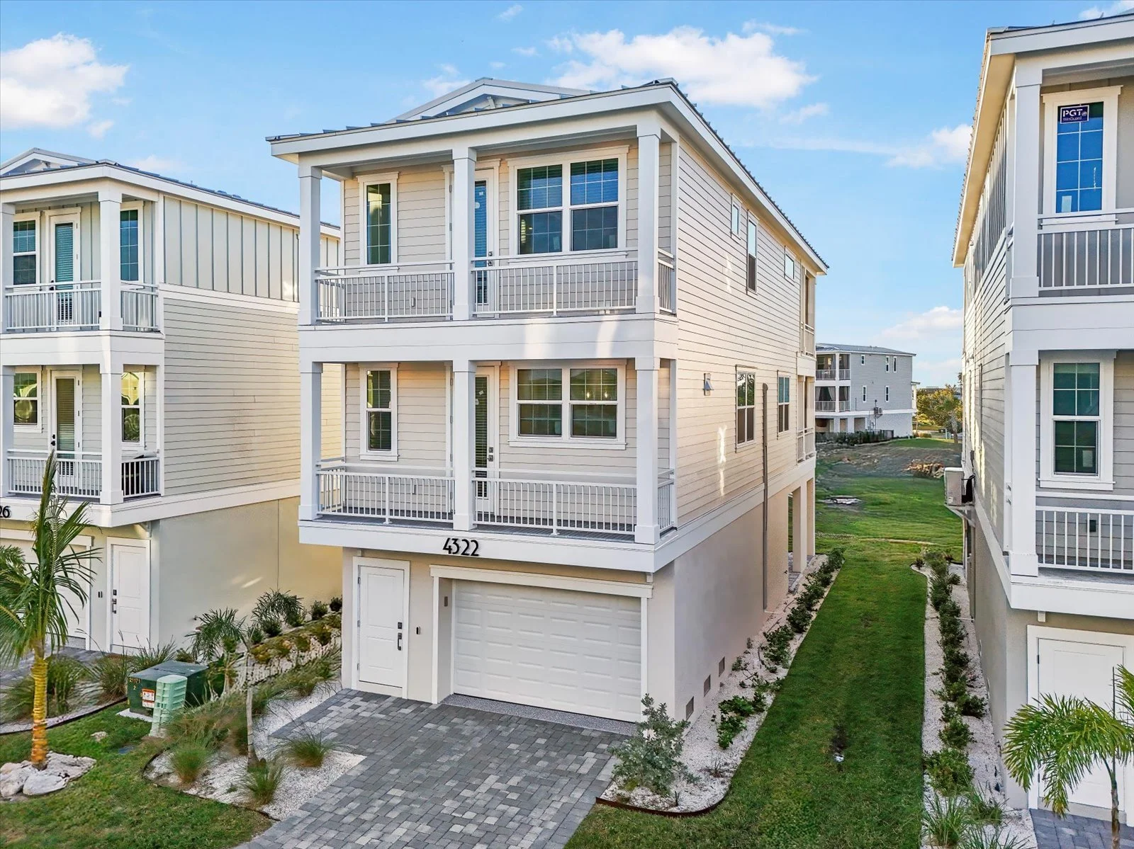 Three-story modern beach house with white siding, multiple balconies, and a garage on the ground level, situated in a landscaped neighborhood with similar houses and green lawns.