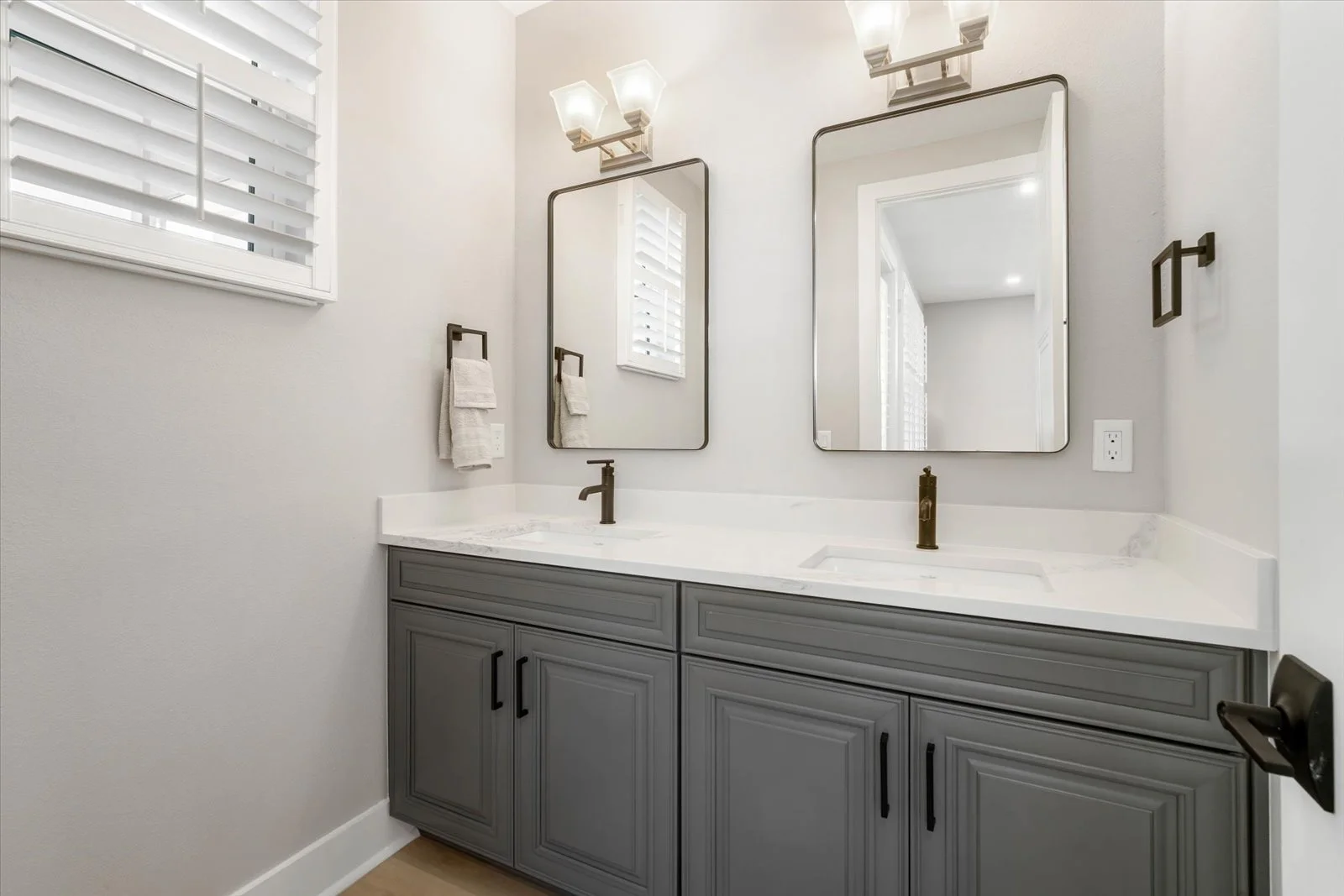 A modern bathroom vanity with a dual sink, gray cabinetry, white marble countertop, two rectangular mirrors, and two wall-mounted light fixtures with frosted glass shades. A window with white shutters is on the left wall, and a towel ring with a fold