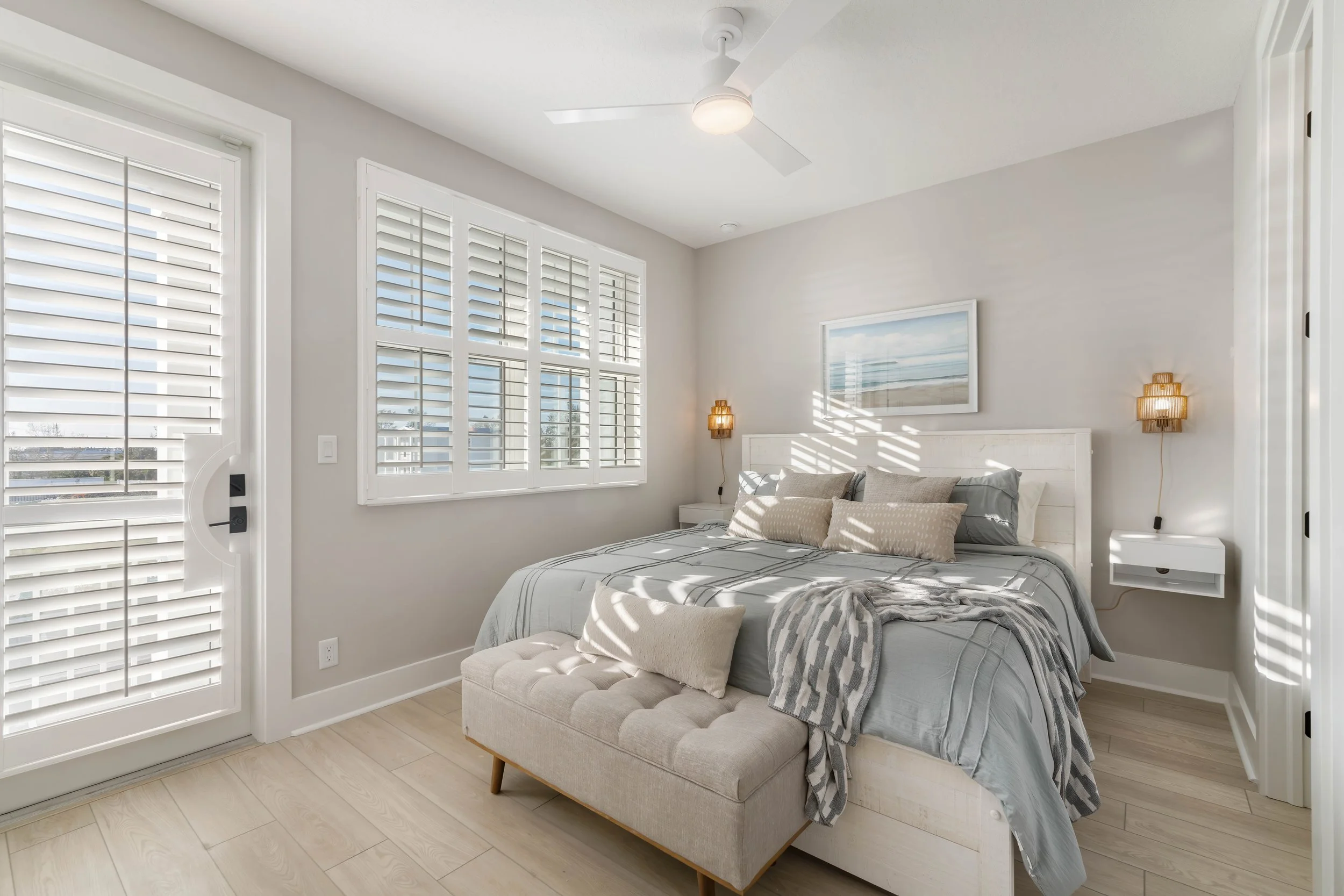 Bright bedroom with a white bed, beige pillows, and a gray blanket, illuminated by sunlight through white shutters.