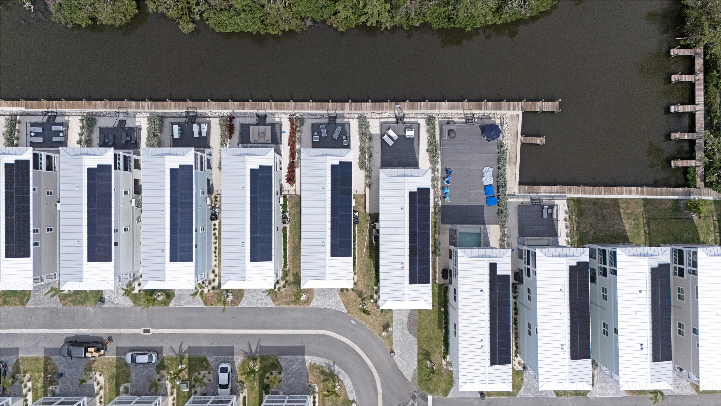 An aerial view of a residential neighborhood with multiple white houses, each with solar panels on the roofs, situated along a curved street. yards with trees and grass, and a waterway runs parallel to the houses at the top of the image. Some houses have driveways with parked cars, and there are wooden docks extending into the waterway.
