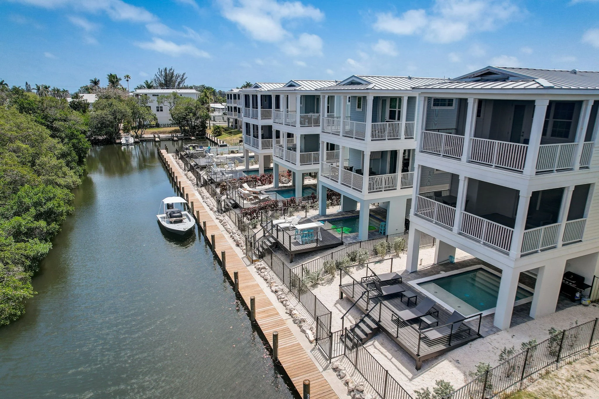 View of modern white multi-story residential buildings with balconies along a canal, featuring patios, hot tubs, lounge chairs, and a dock with boats under a blue sky.