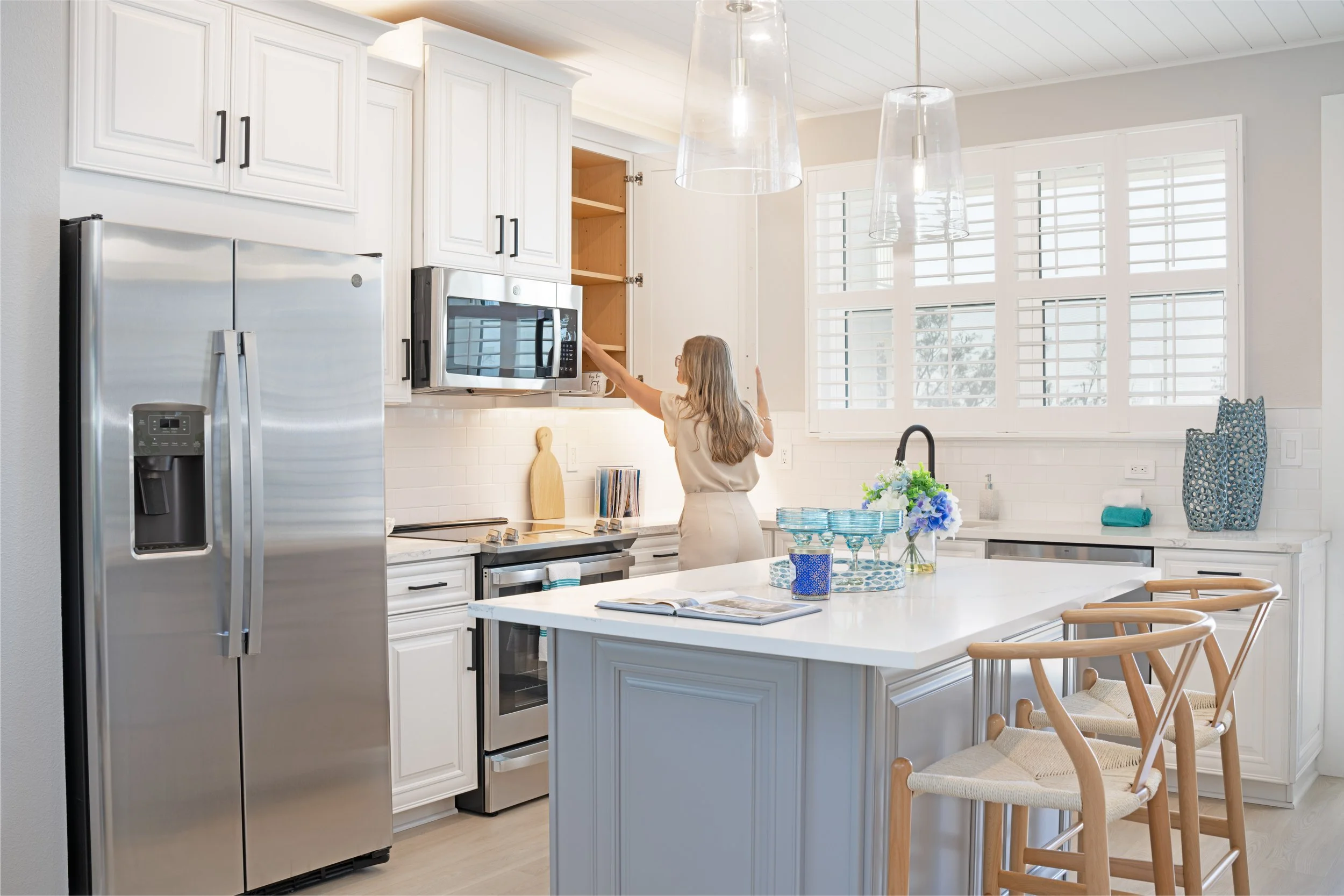 A woman is reaching into a kitchen cabinet above the microwave, with white cabinets, a stainless steel refrigerator, a kitchen island with a book and decorative items, and large windows with white shutters.