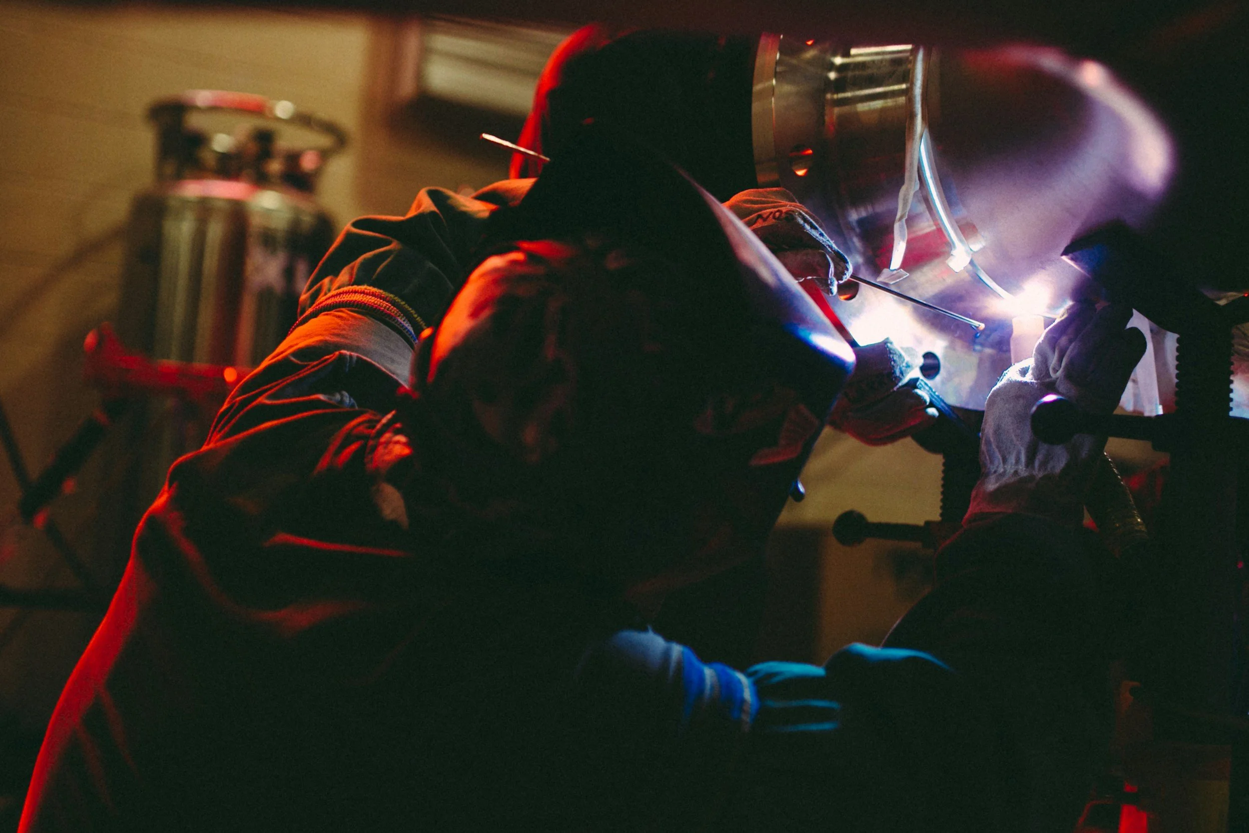 Person welding metal, wearing protective helmet and gloves, with bright sparks and glow from welding process.