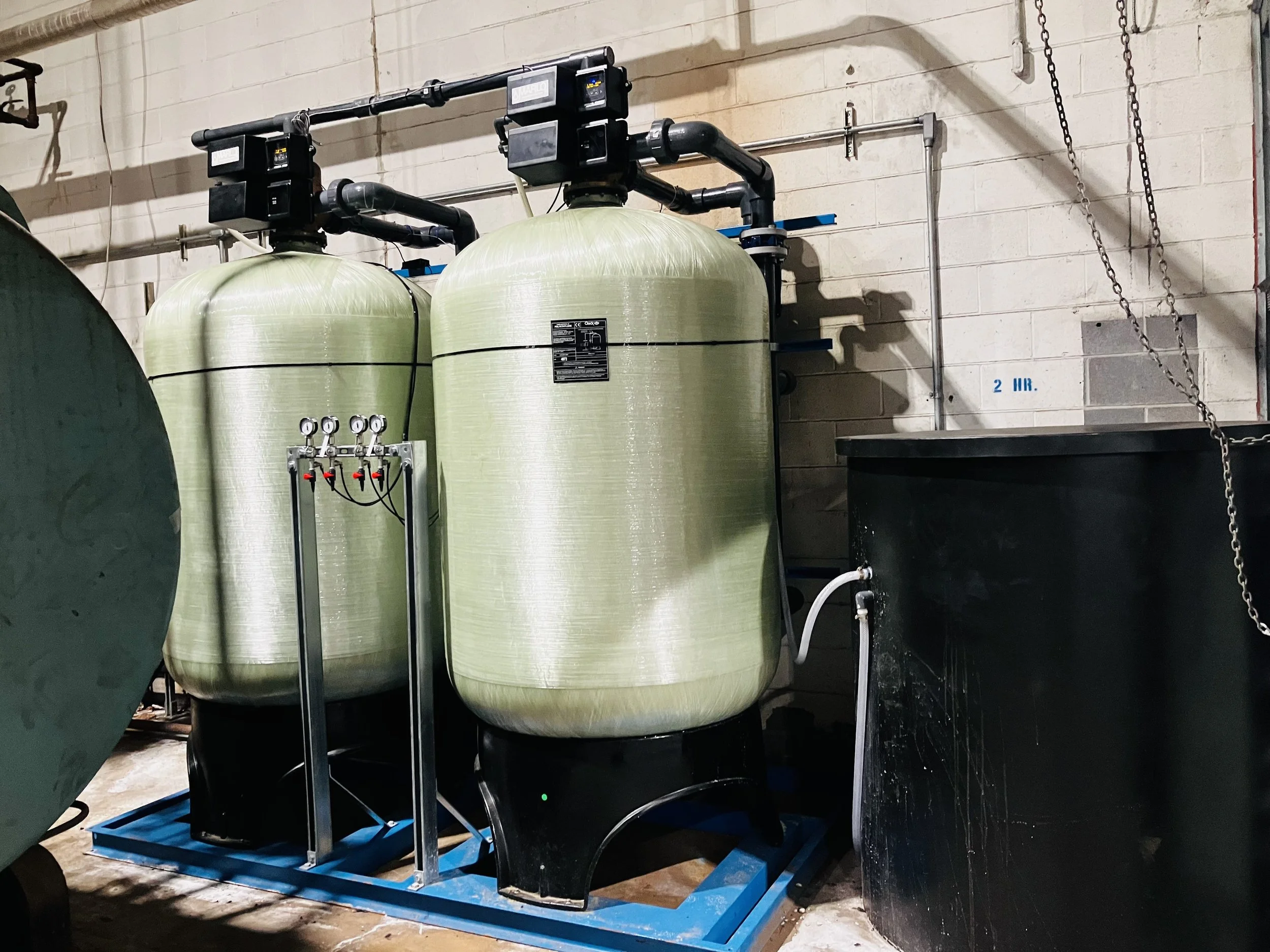 Two large green water filtration tanks with black bases, connected by black pipes, standing on a blue metal frame, next to a black container in an industrial setting.