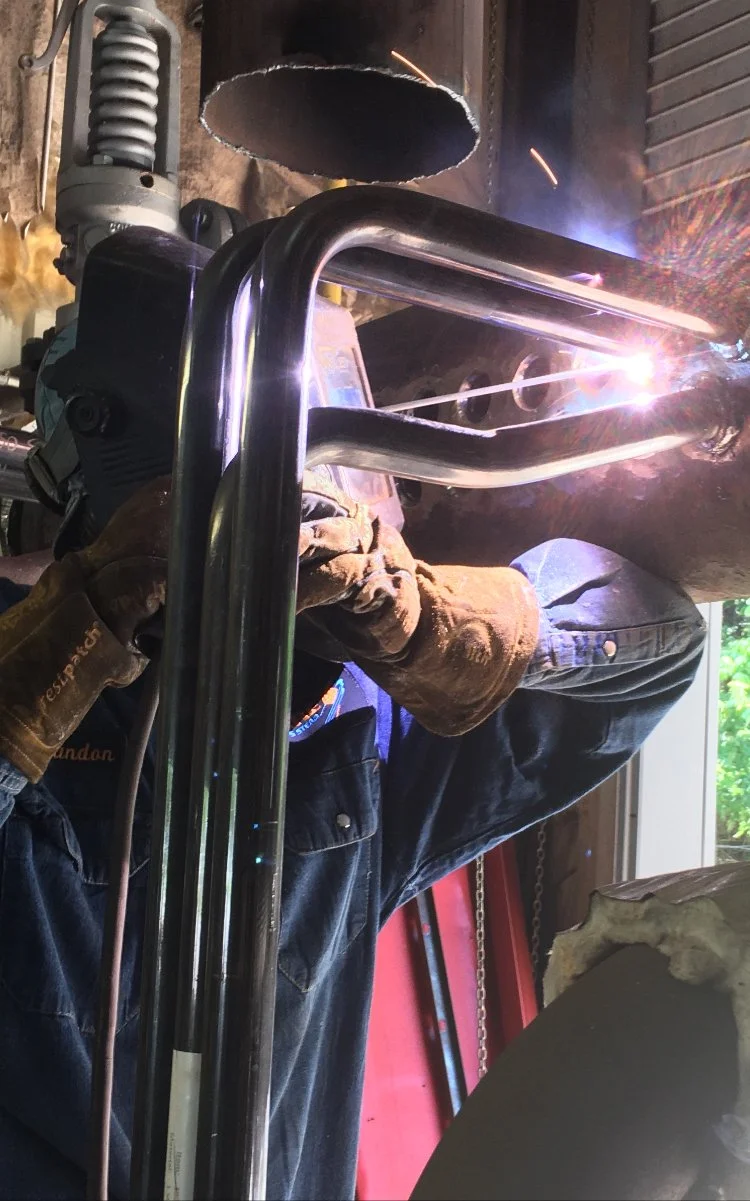 A person welding metal pipes in a workshop, sparks flying from the welding process, wearing protective gloves and a safety jacket.