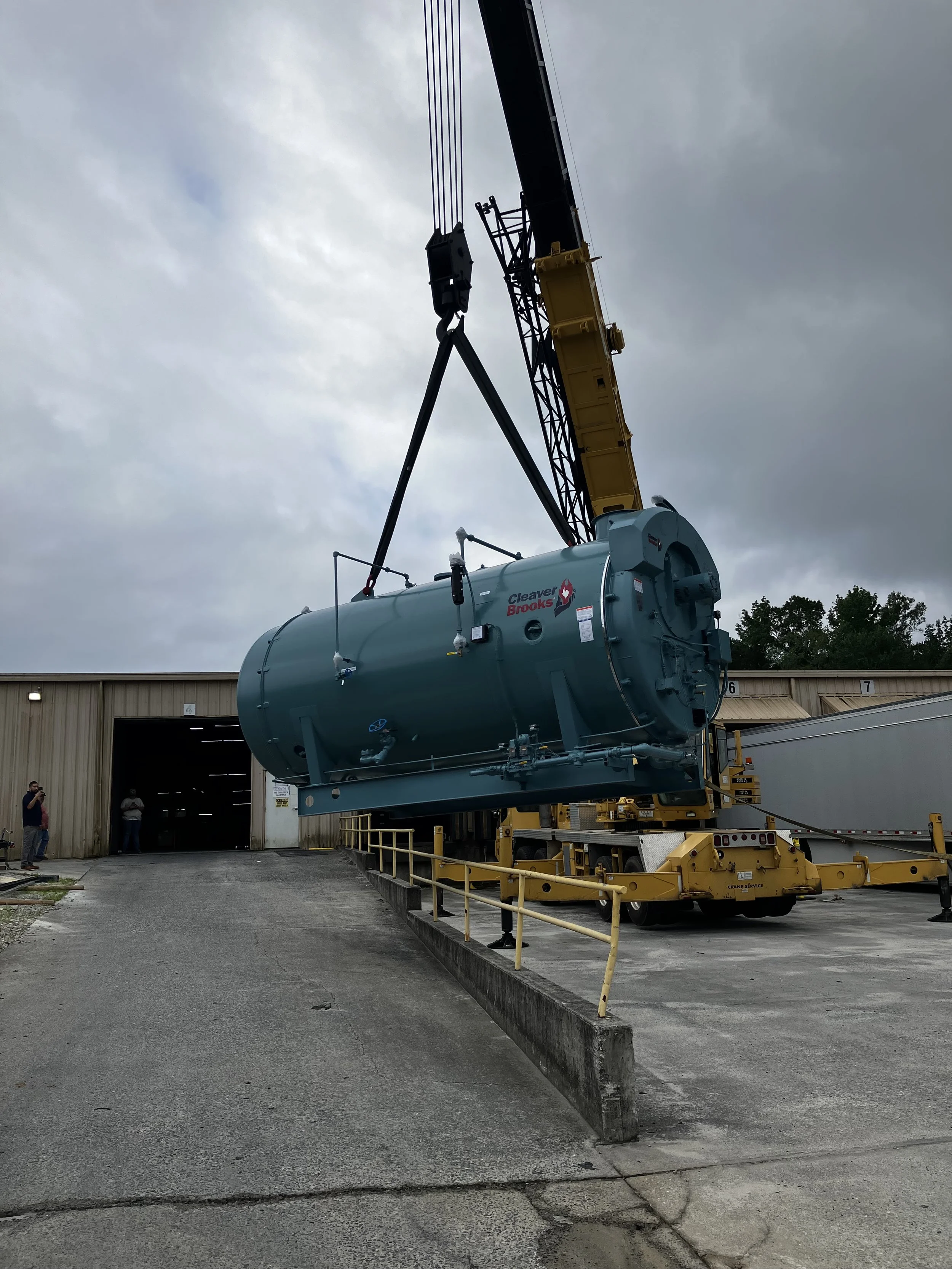 A large industrial crane lifting a blue tank labeled 'Cleaver Brooks' outside a warehouse.