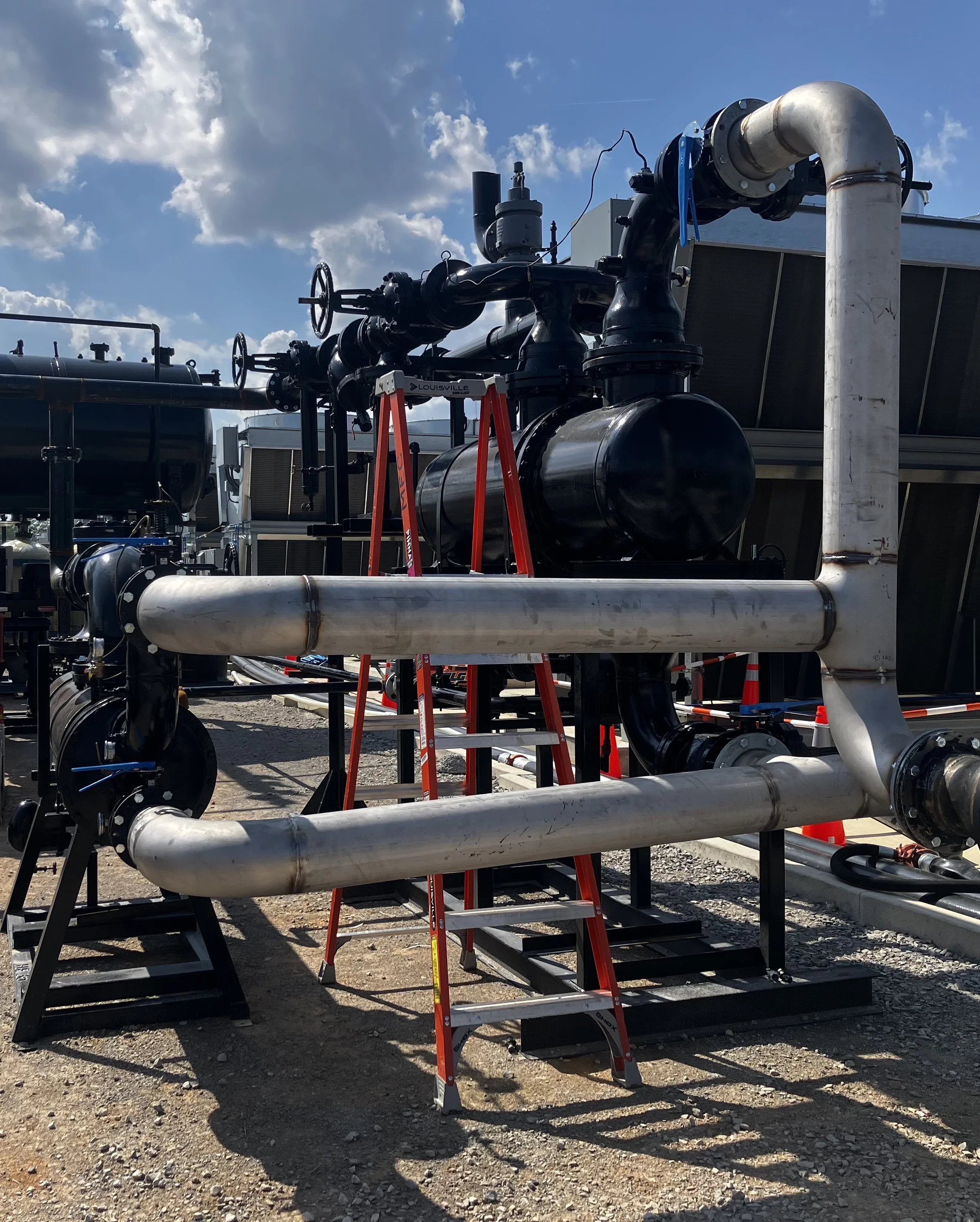 Industrial piping and mechanical equipment on outdoor site, with ladders and bright partly cloudy sky.