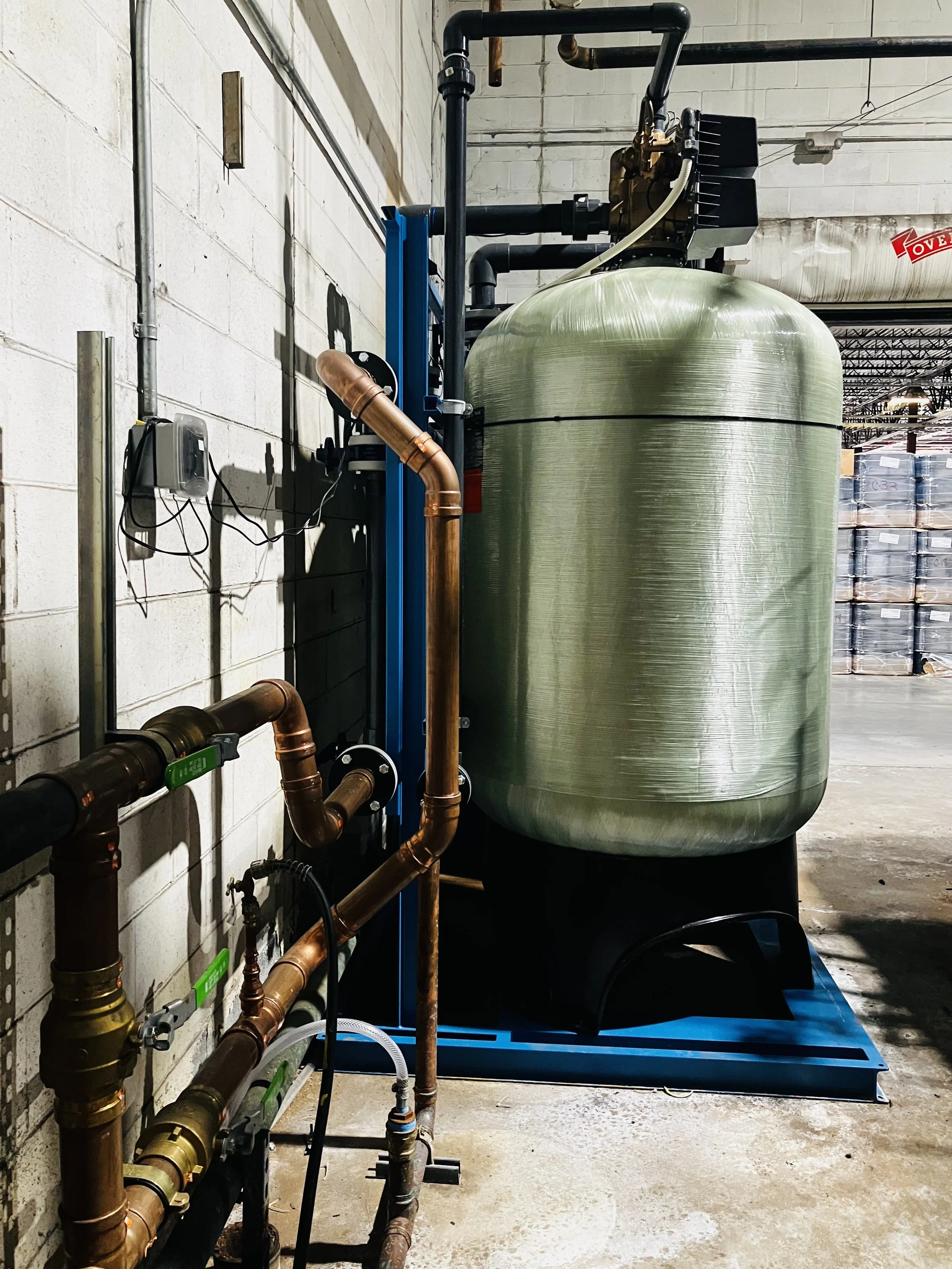 Industrial water treatment system with a large green tank, copper pipes, and control components in a warehouse.